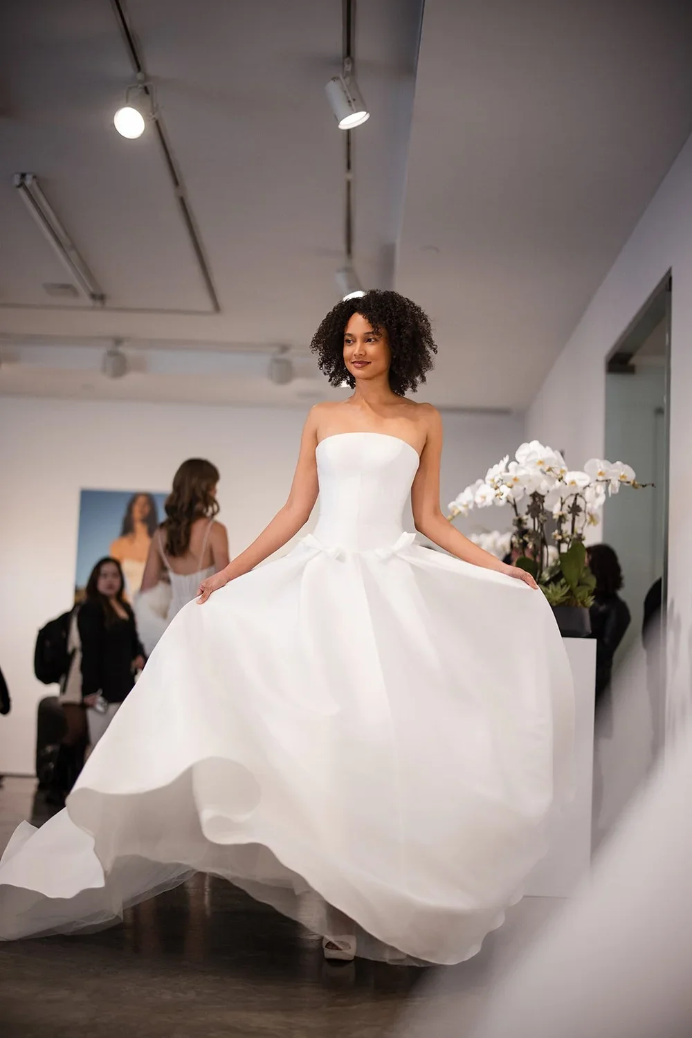 A woman trying on a white strapless ball gown wedding dress in a bridal boutique, holding the sides of her dress, with flower arrangements nearby and other women in the background.