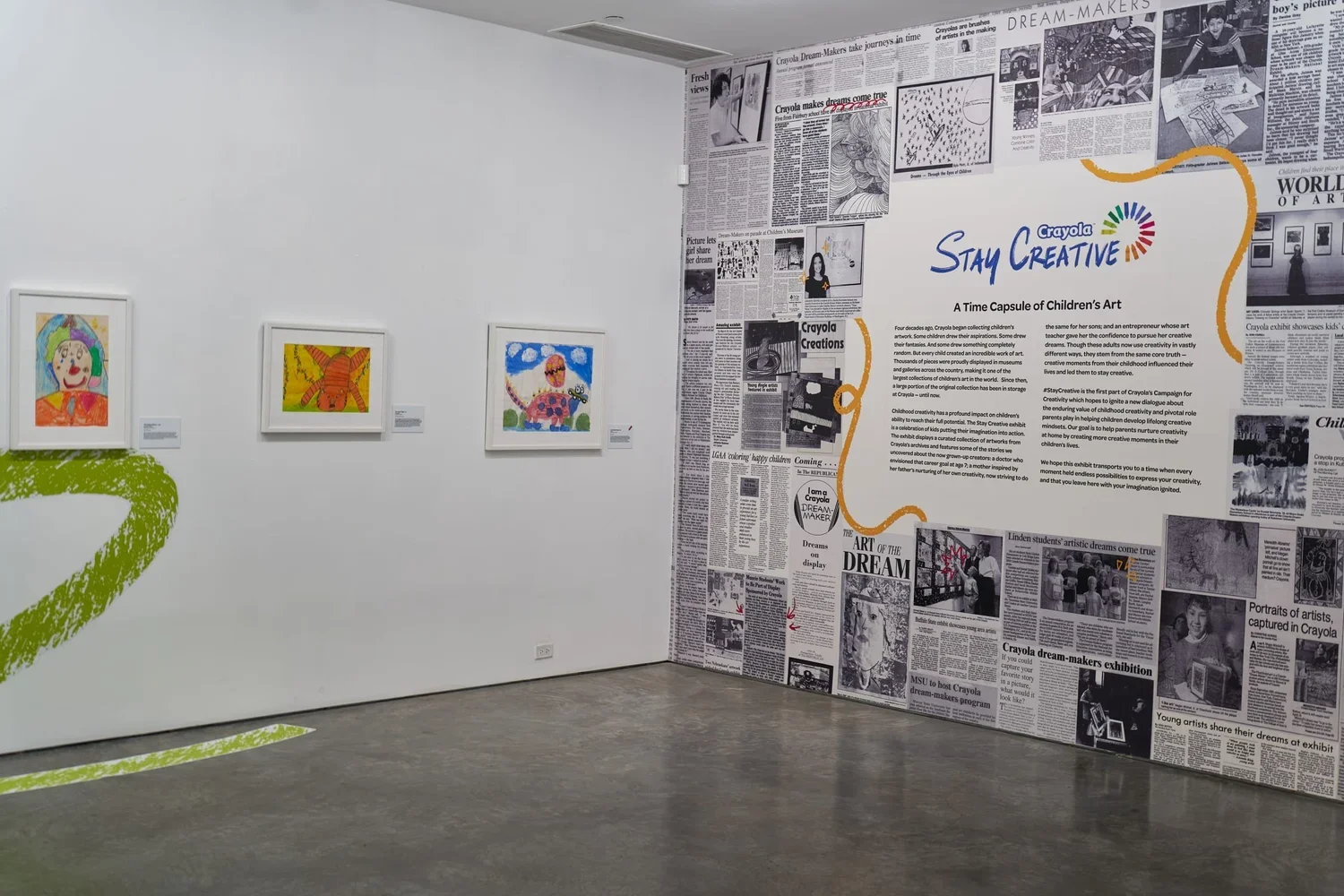 An art exhibition featuring children's colorful drawings on the wall with three framed artworks displayed on a white wall and a large collage of newspaper clippings about children and art on the adjacent wall. The right wall has text about Crayola's 