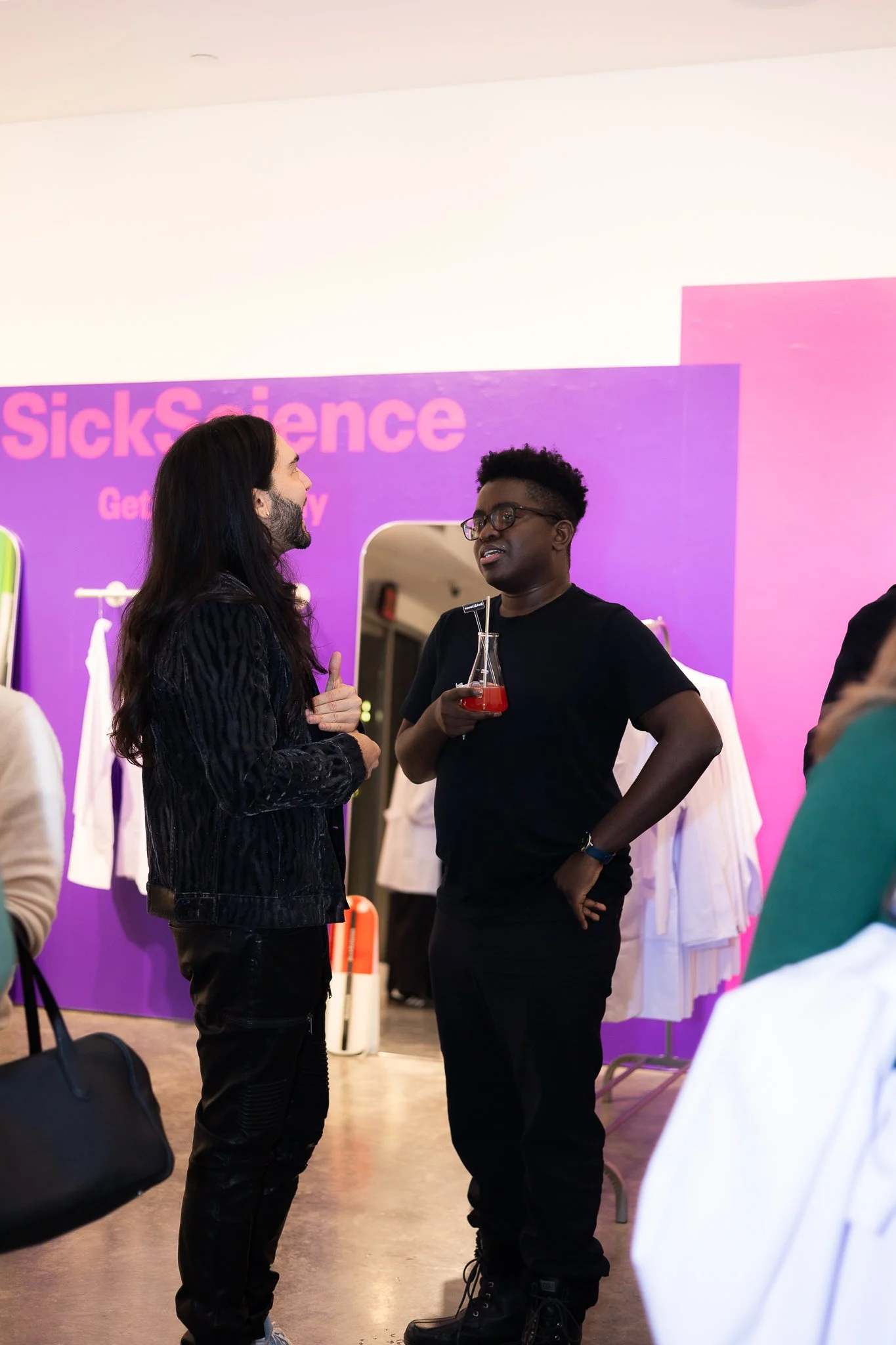 Two people engaged in conversation at an event with a purple and pink backdrop that reads 'SickScience Get'. One person has long dark hair, wears a black jacket, and is gesturing towards the other individual with a beverage in hand. The other person 