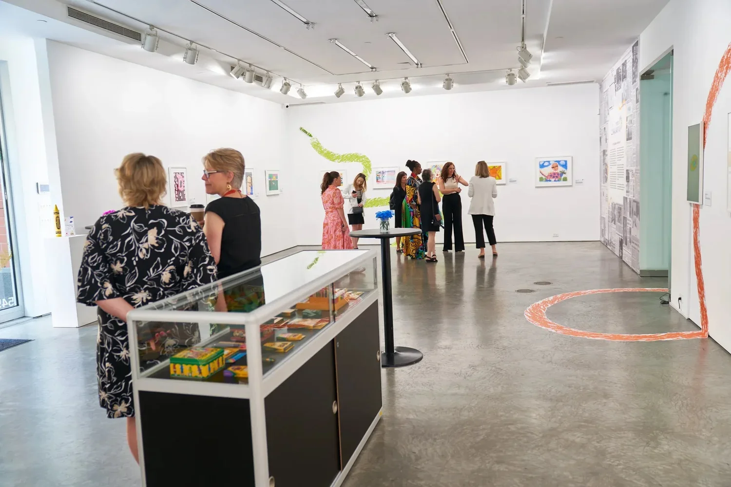 People viewing artwork in a gallery with white walls and colorful paintings. Two women are near the front on the left, and a group of people in the background are observing the art. There are track lights on the ceiling and a display case with books 