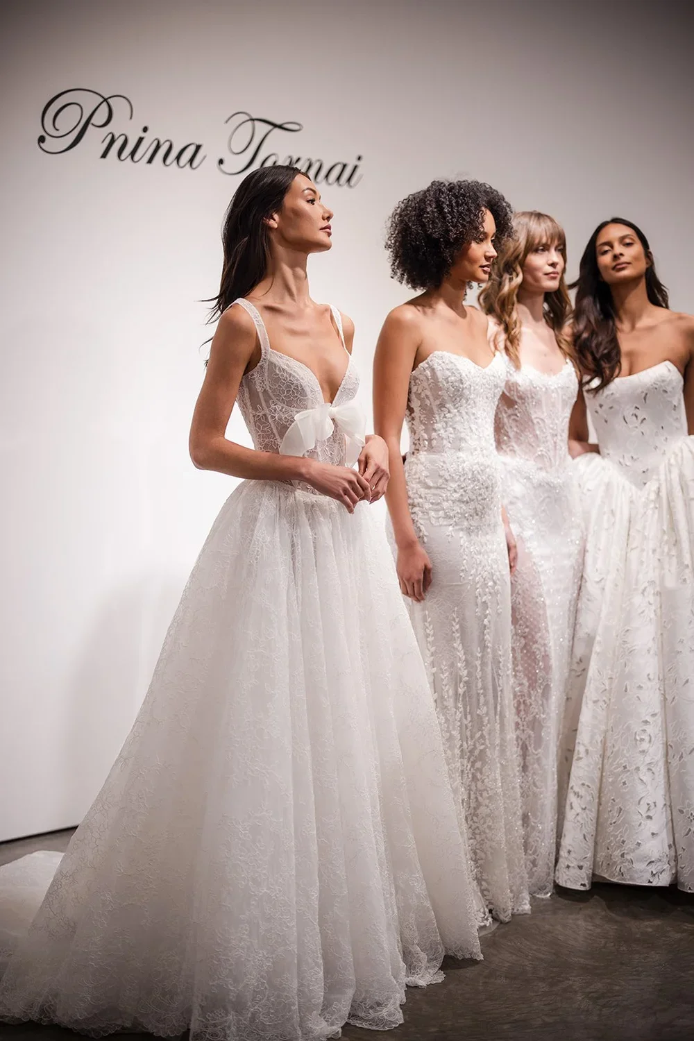 Four women in wedding dresses standing in a line at a bridal fashion show or boutique.