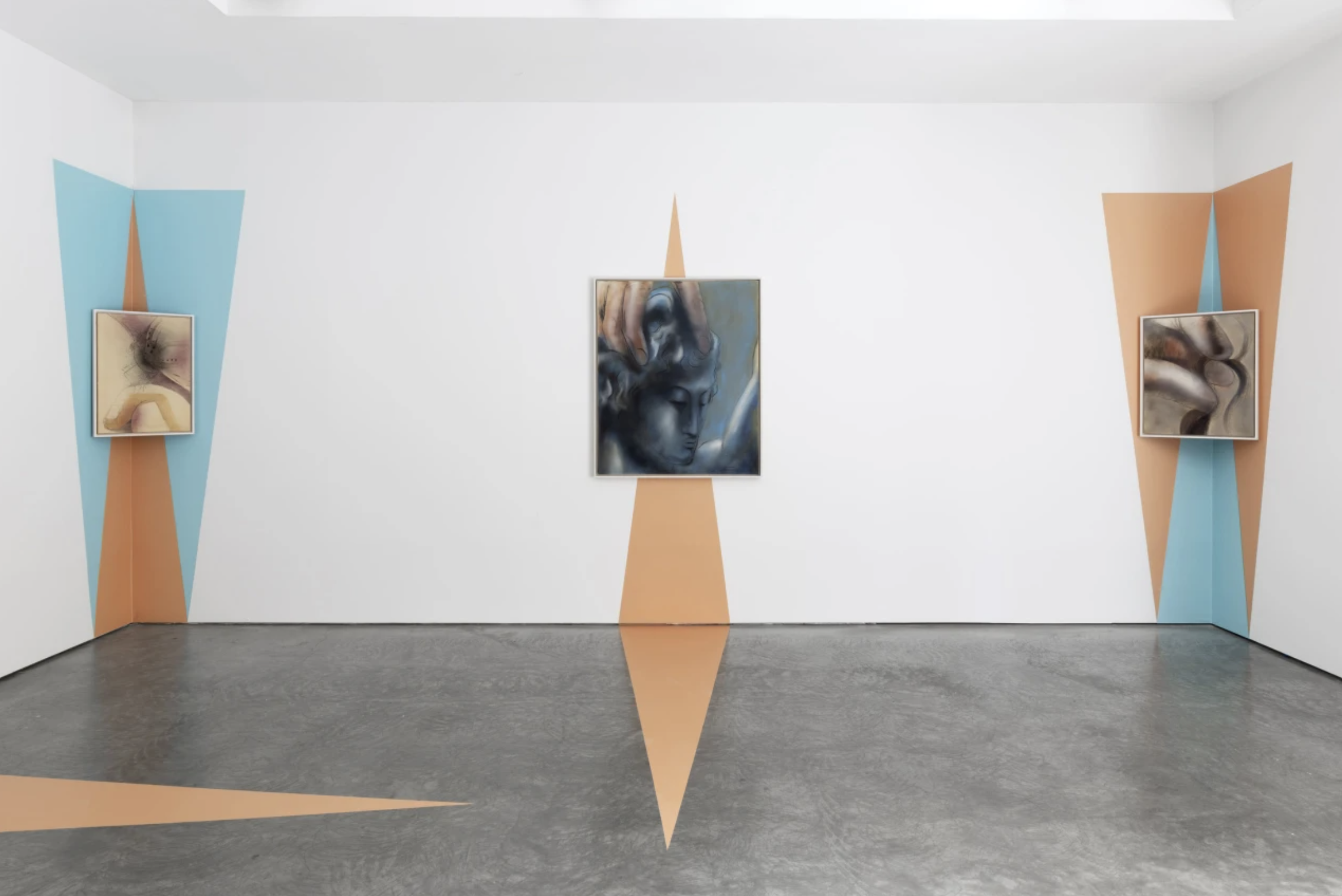 A contemporary art gallery display with three abstract paintings hung on a white wall. The paintings are framed and mounted on colorful geometric backgrounds painted on the wall in shades of blue and orange. The flooring is dark gray concrete with ma