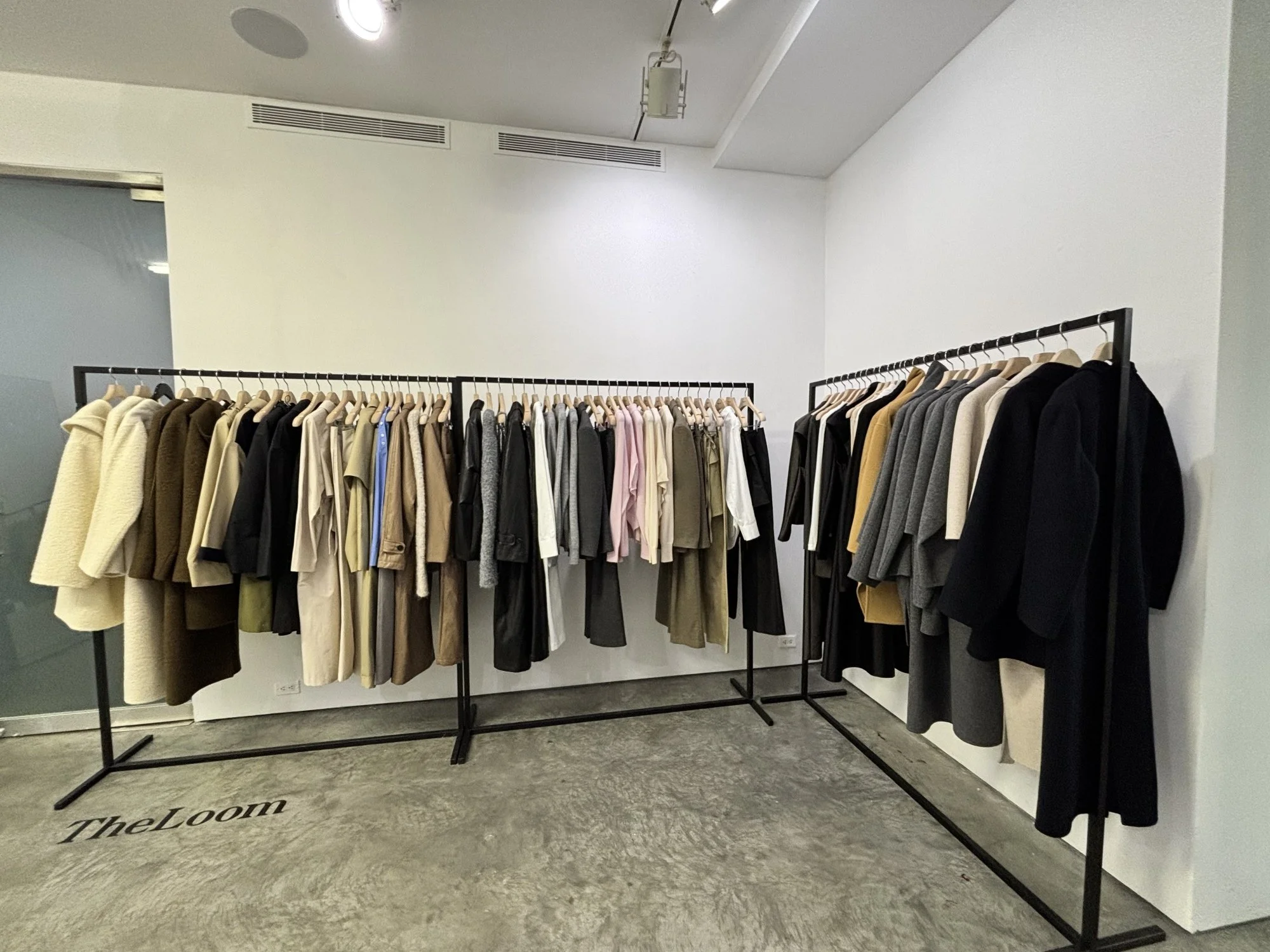 Clothing racks with beige, black, gray, pink, and white coats in a store called TheLoom.