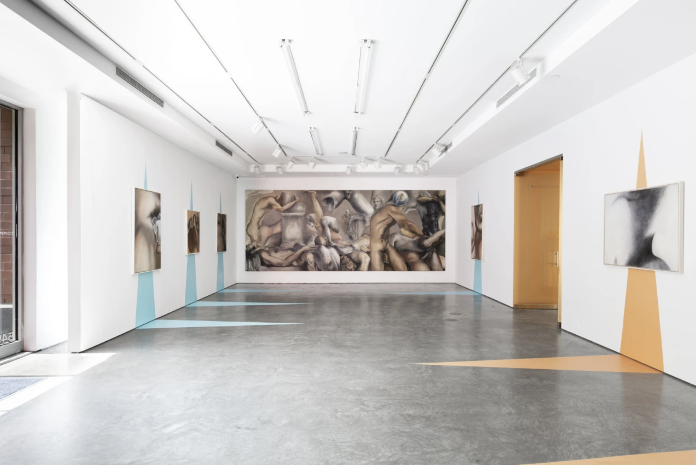 Empty art gallery with white walls, featuring several modern paintings, including a large mural of intertwined human figures at the back, and geometric painted shapes on the lower walls.