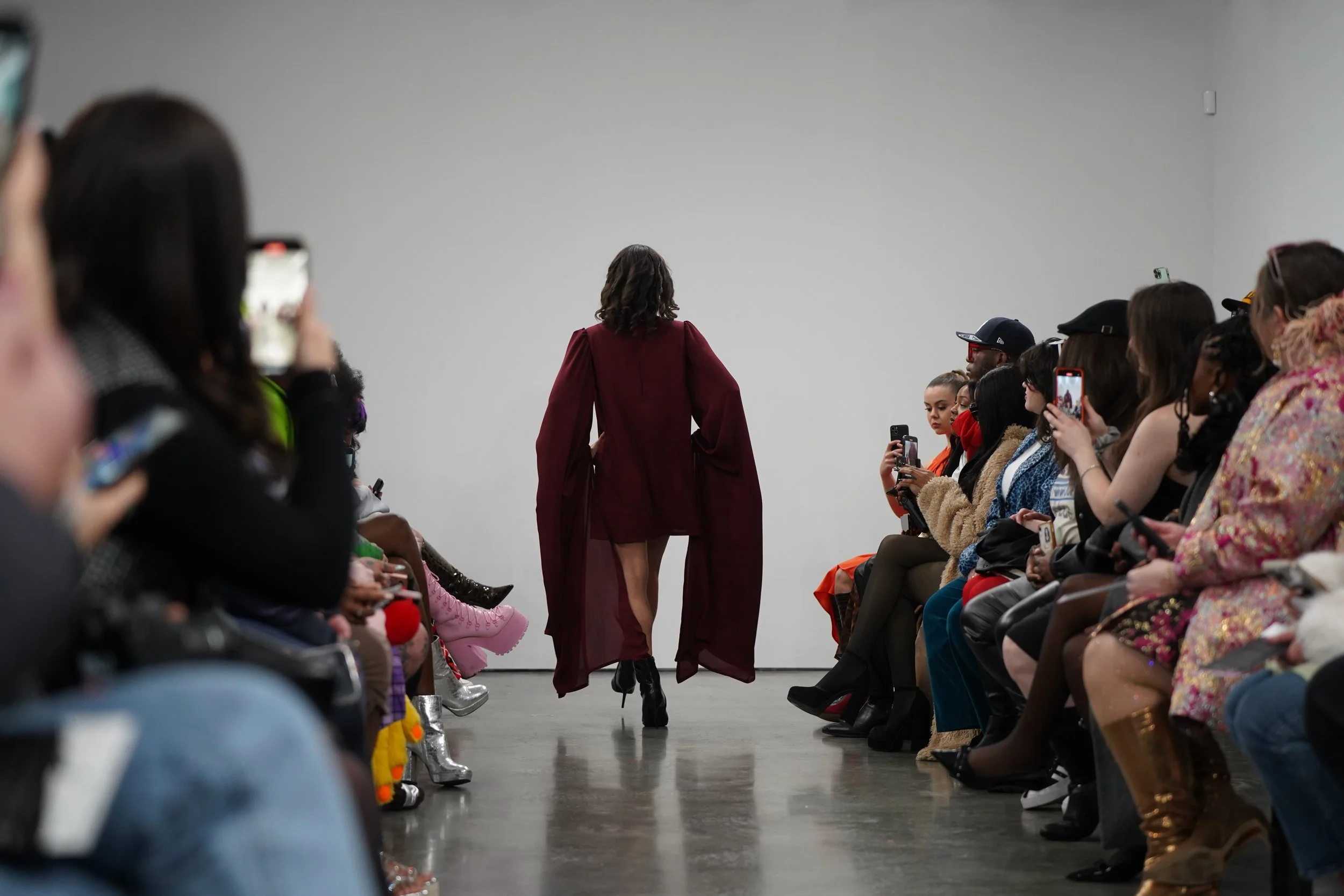 A model walking down a runway during a fashion show, wearing a maroon outfit with a high slit, surrounded by seated audience members taking photos.