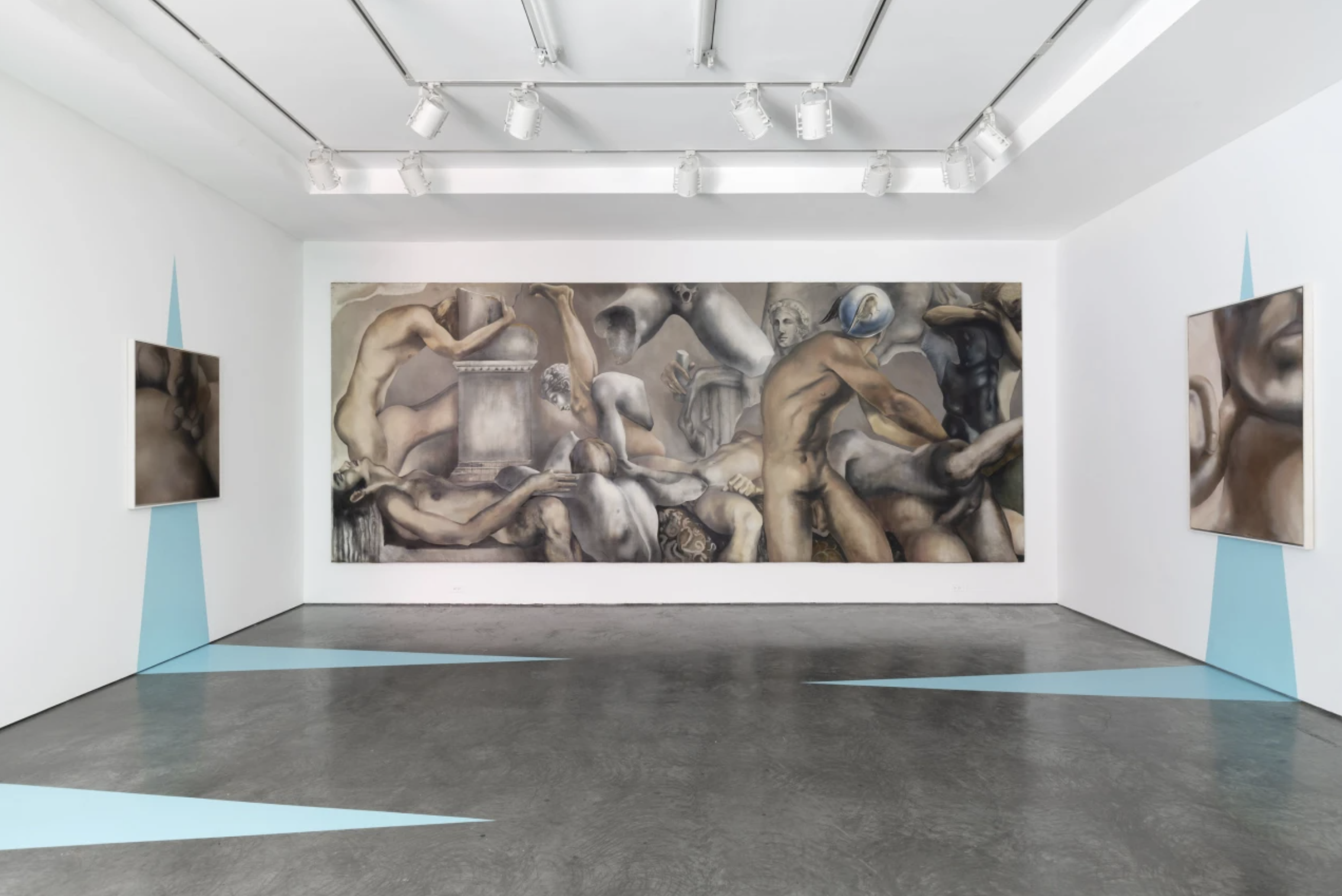 An art gallery with white walls, showcasing three paintings. The central large painting depicts numerous nude human figures with muscular builds in a complex arrangement, some watching and others engaged in various poses. The two smaller paintings on