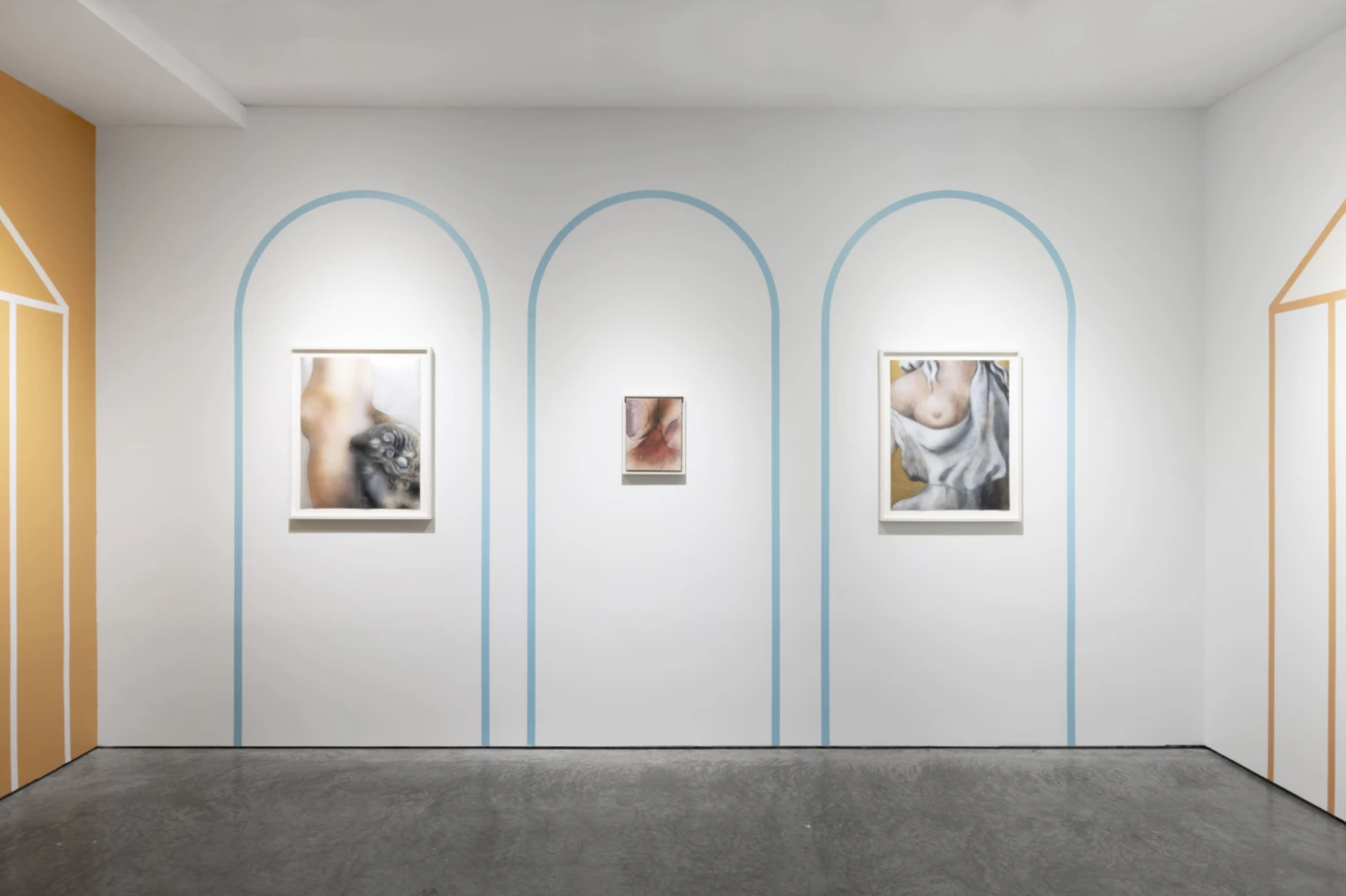 Art gallery with three framed artworks on white wall, decorative arches outlined in blue and orange, polished concrete floor.