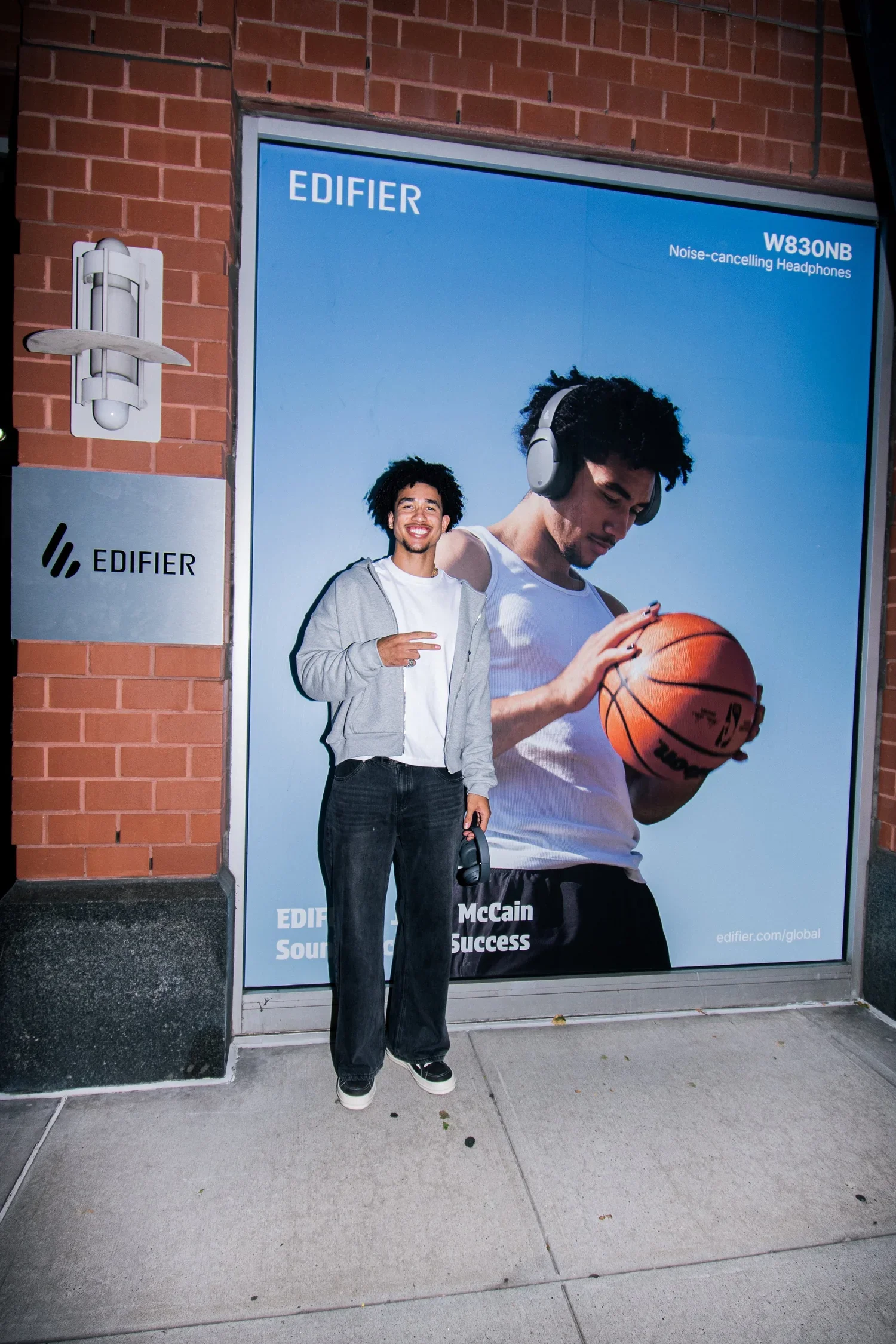 A young man is standing outside a brick building, holding a pair of headphones in his right hand and smiling at the camera. Behind him is a large advertisement featuring a person wearing headphones and holding a basketball.
