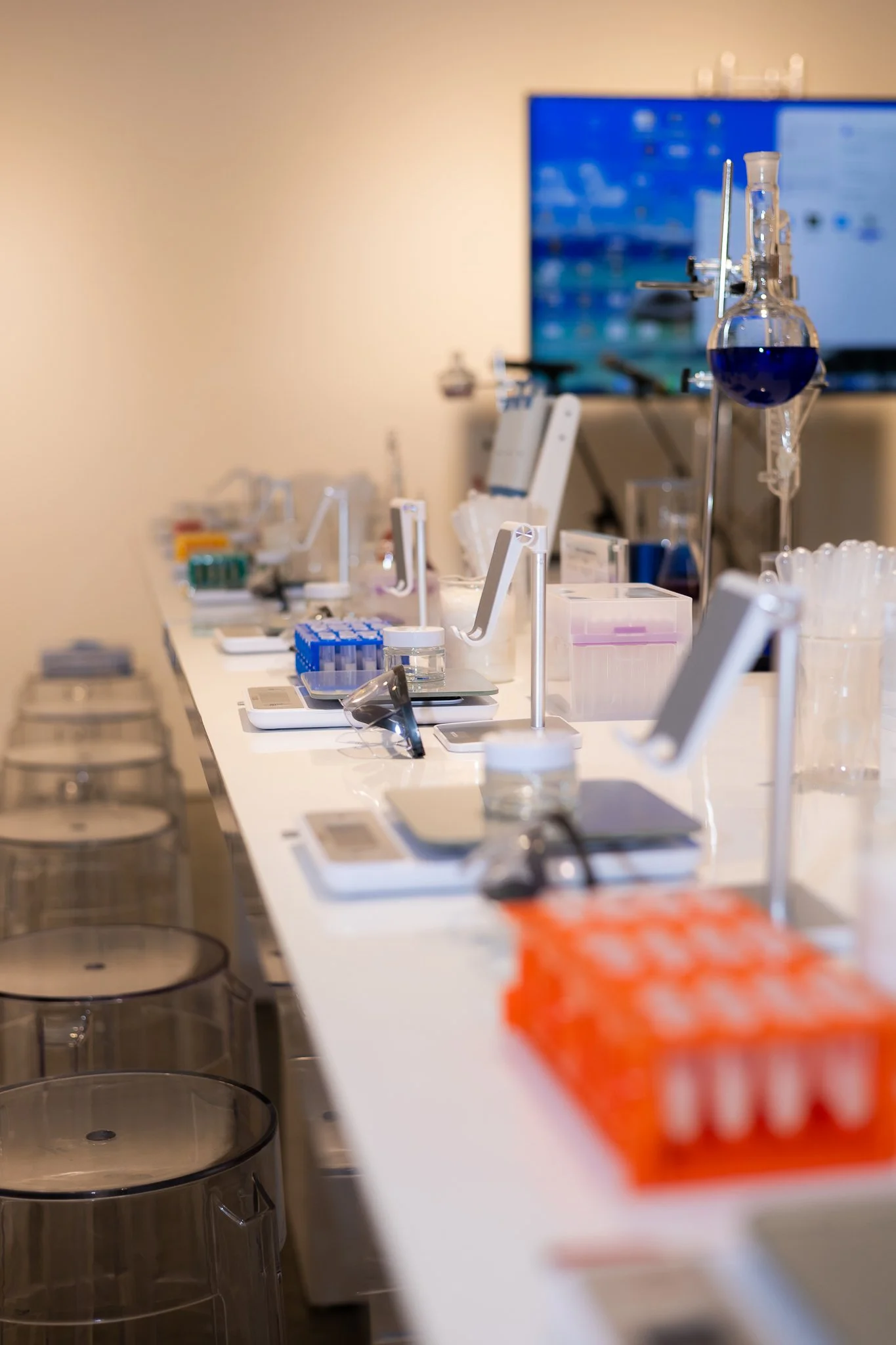 Laboratory with scientific equipment including test tubes, scales, and containers on a white counter, with a large screen displaying data or images in the background.