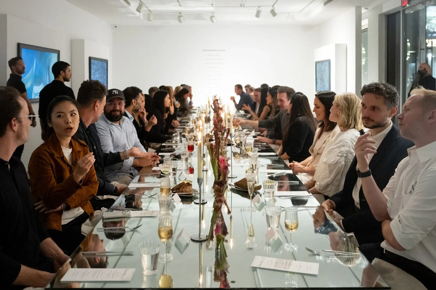 A large group of diverse people sitting at a long, glass-topped table in a modern, well-lit dining setting, engaged in conversation and enjoying a meal with wine and dishes, decorated with flowers and candles.