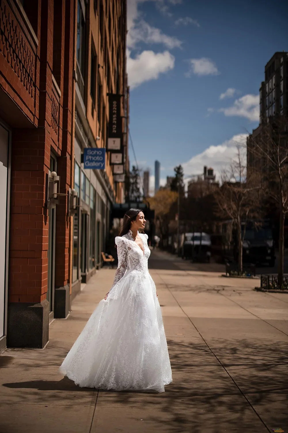 A woman in a white wedding dress standing on a city sidewalk under a blue sky with clouds.