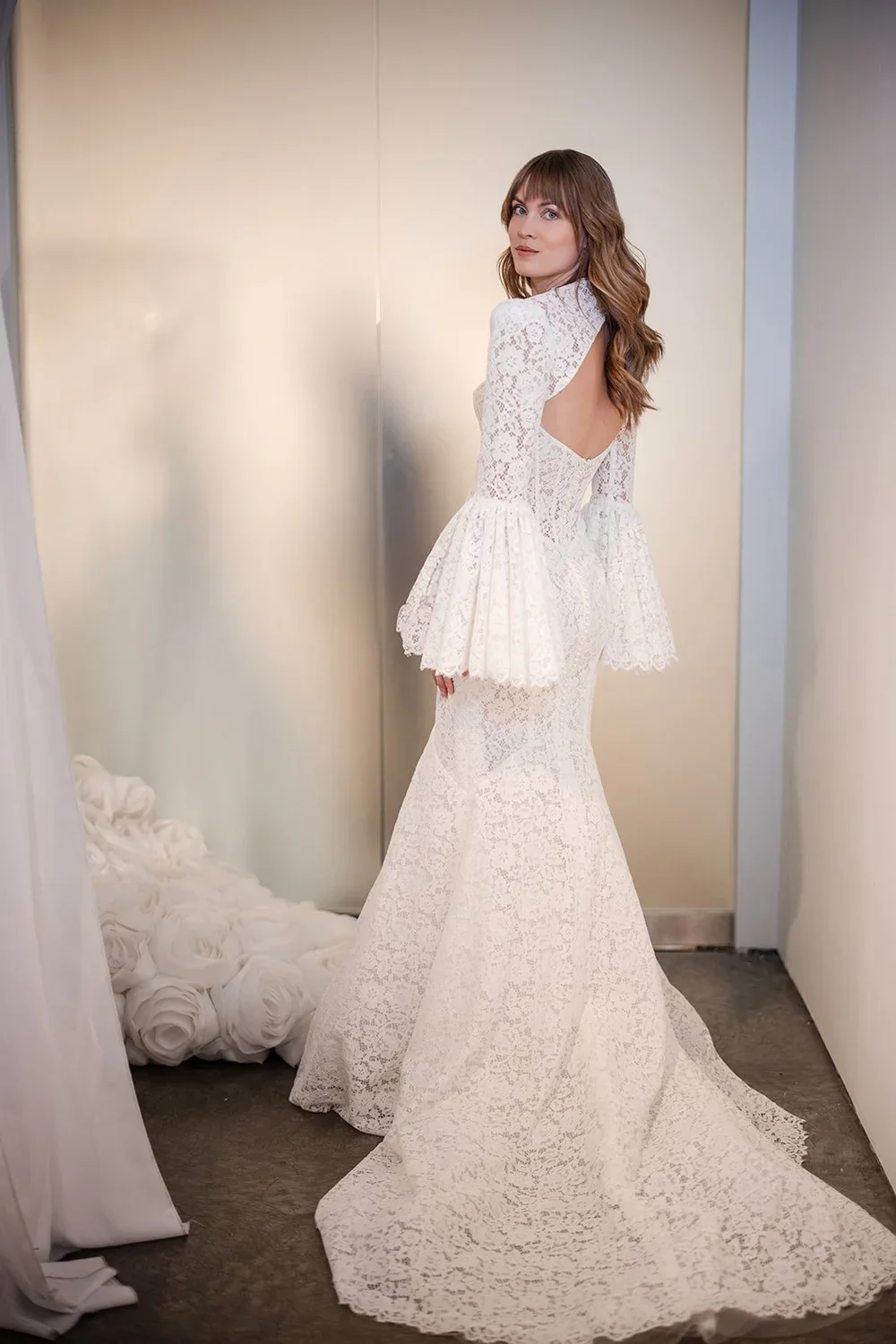 A woman in a white lace wedding dress with open back and flared sleeves, standing in a fitting room with a silver wall and white rosette decor on the floor.
