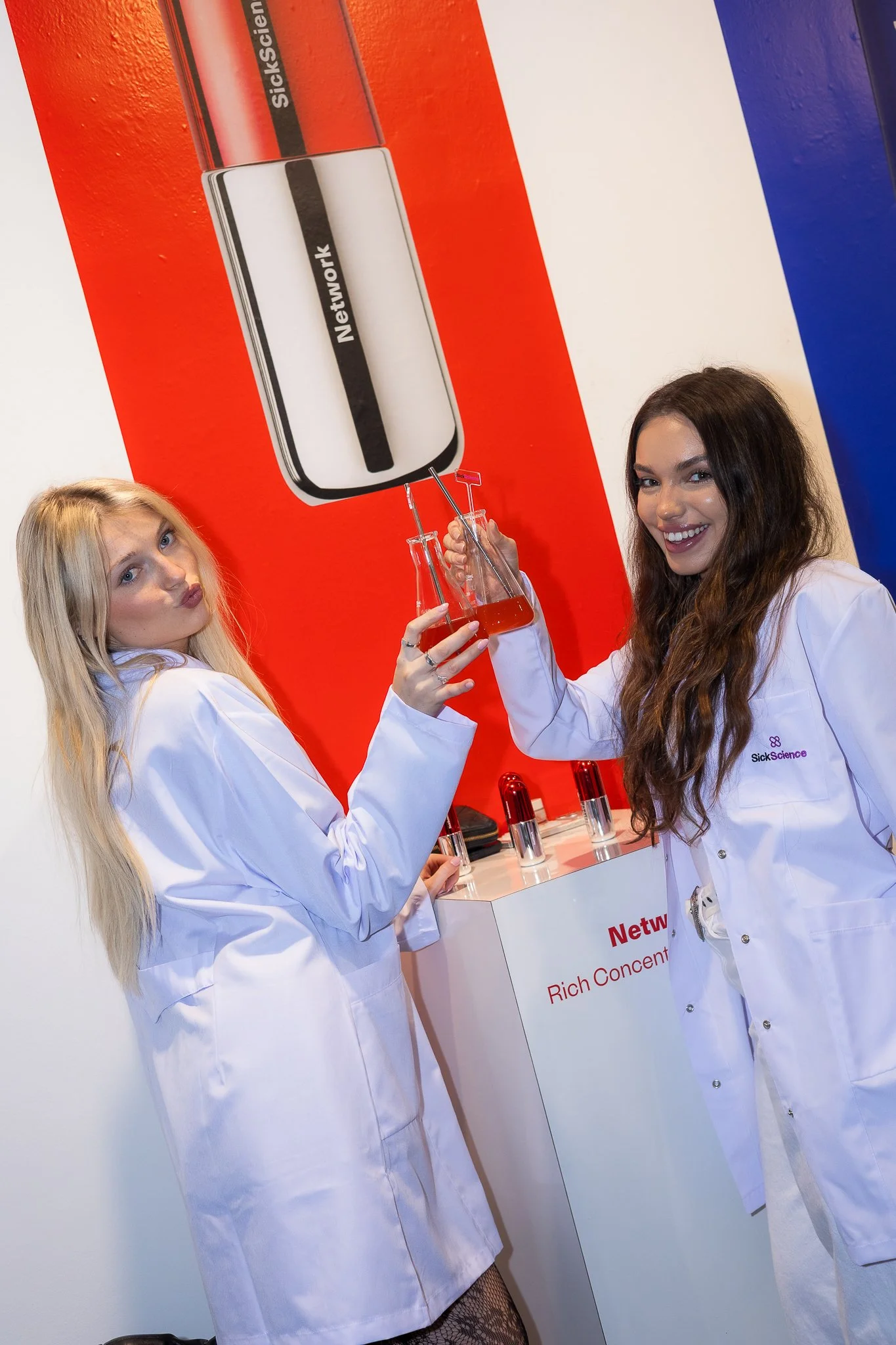 Two women in white lab coats holding test tubes with a red liquid, standing in front of a colorful science exhibit with large bottles and lab equipment.