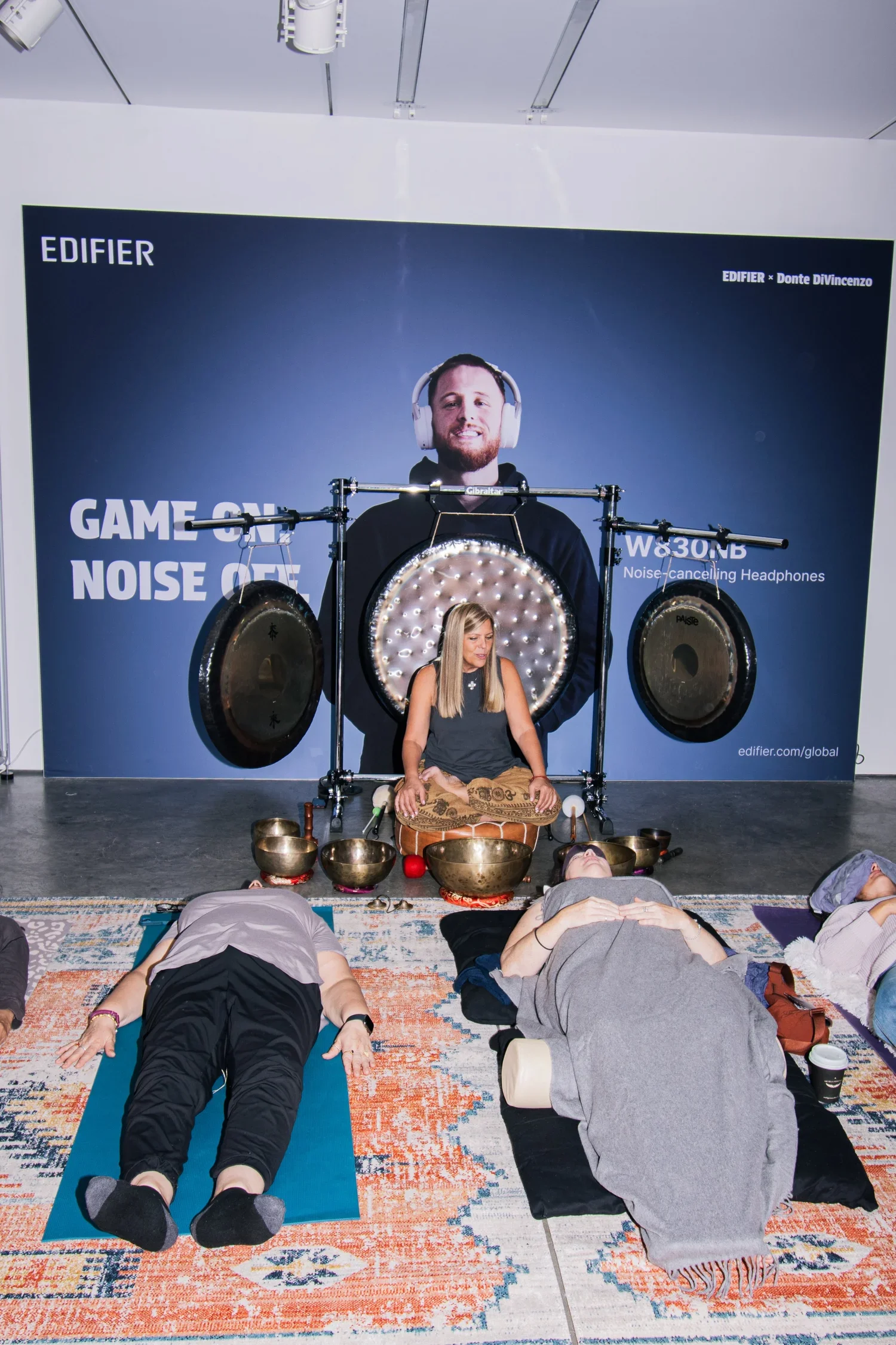 People lying on yoga mats and cushions during a sound healing session with a woman playing Tibetan singing bowls in front of a large advertisement featuring a man wearing headphones and a gong.