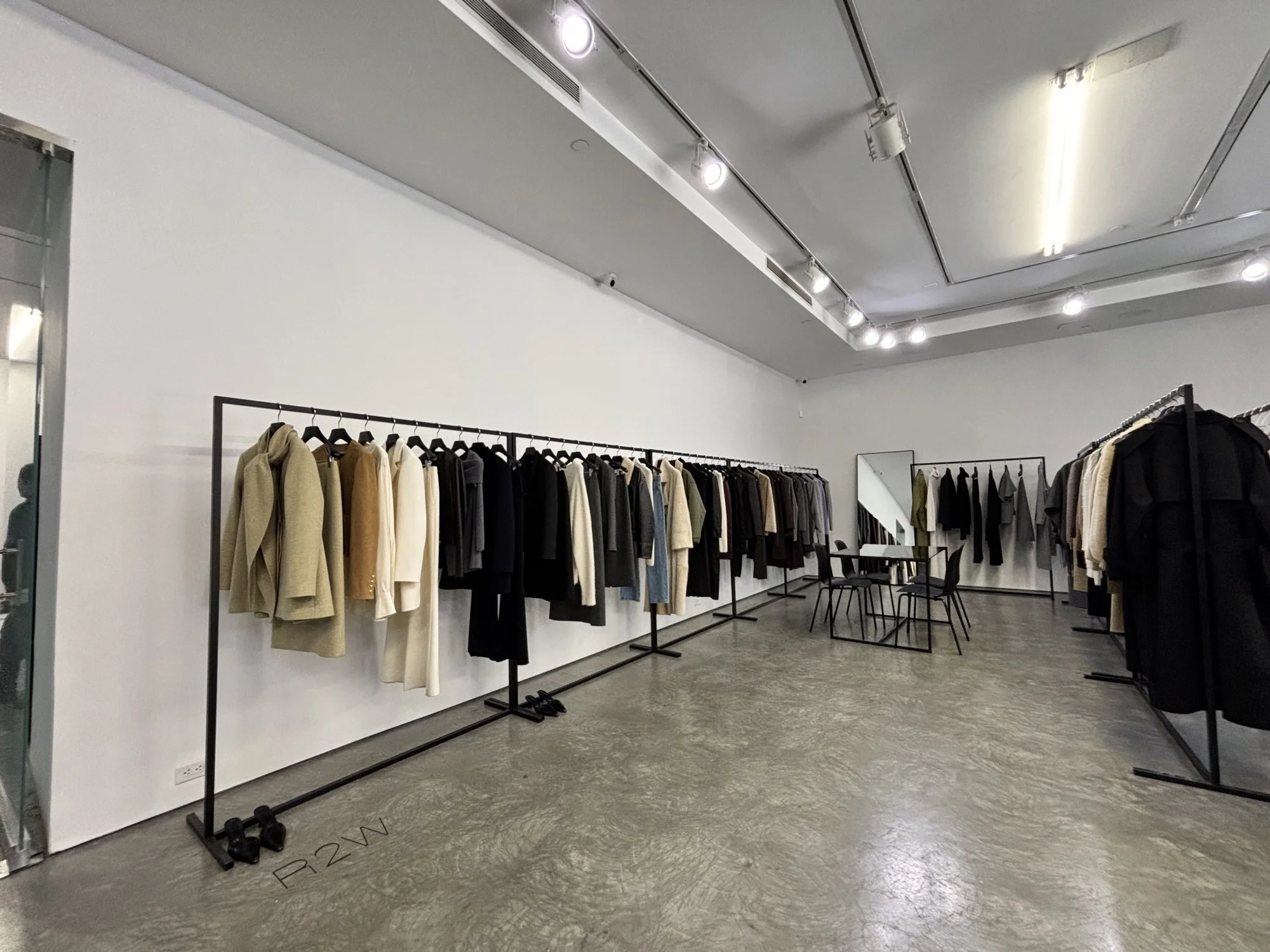 Clothing store interior with racks of neutral-colored coats, a large mirror, and table and chairs.