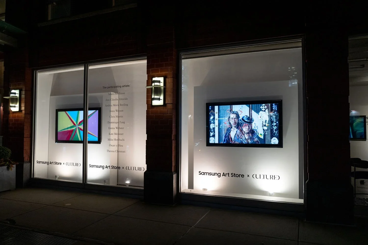 Storefront window display with digital screens showcasing colorful artwork, titled "Samsung Art Store x CULTURE." List of participating artists on the glass, illuminated wall sconces, and dark brick exterior.