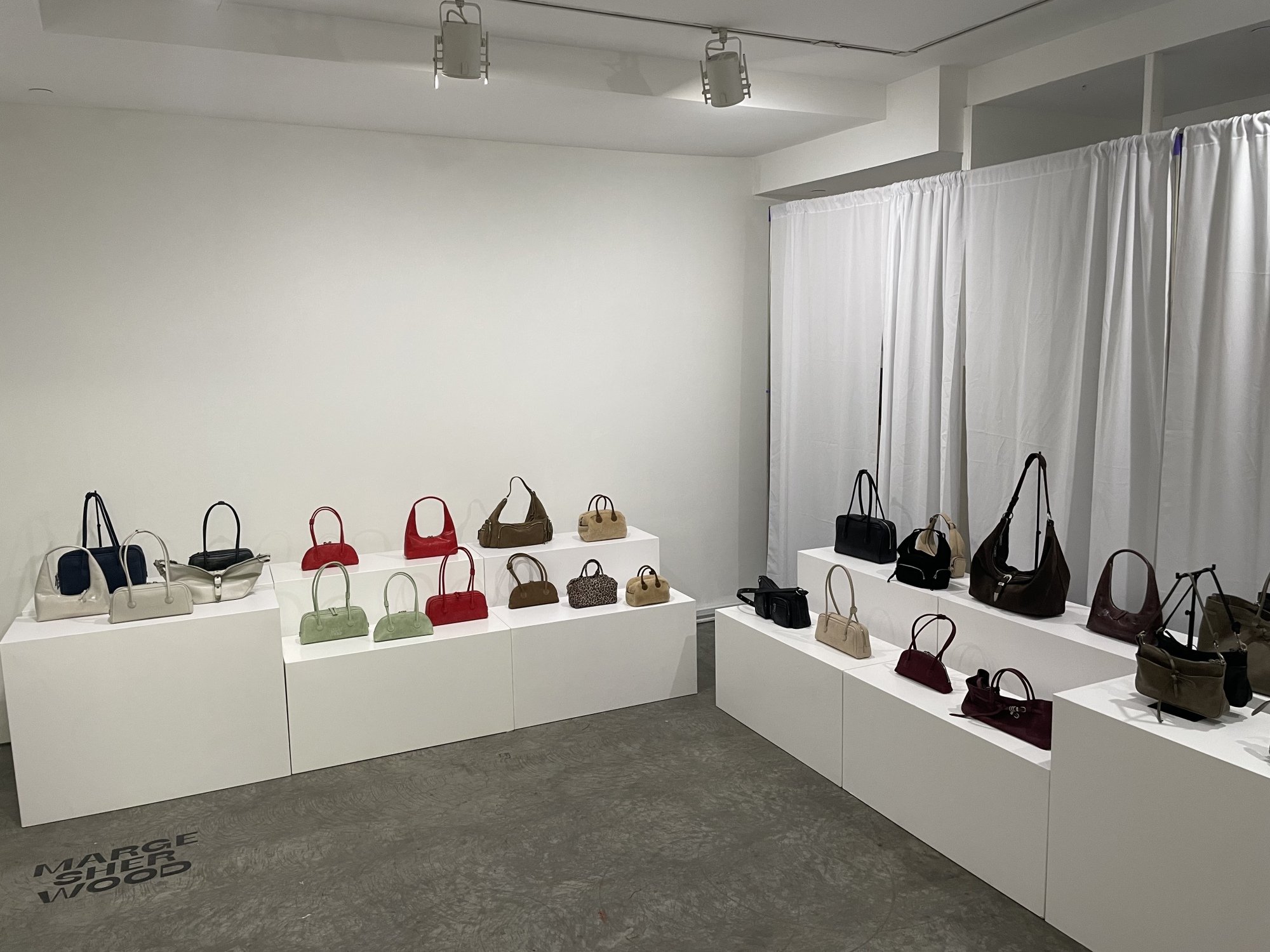 Display of various handbags in a minimalist gallery setting with white walls and pedestal stands, organized by color and style.