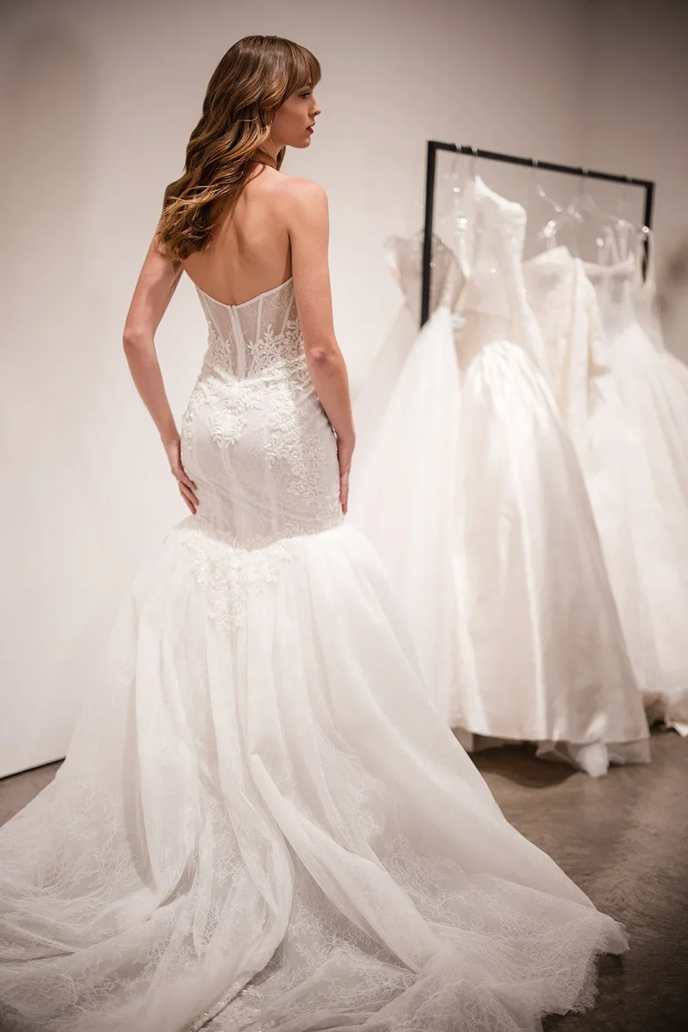 Woman trying on a strapless, lace wedding dress with a mermaid silhouette in a bridal boutique, with several other wedding dresses hanging on a rack in the background.