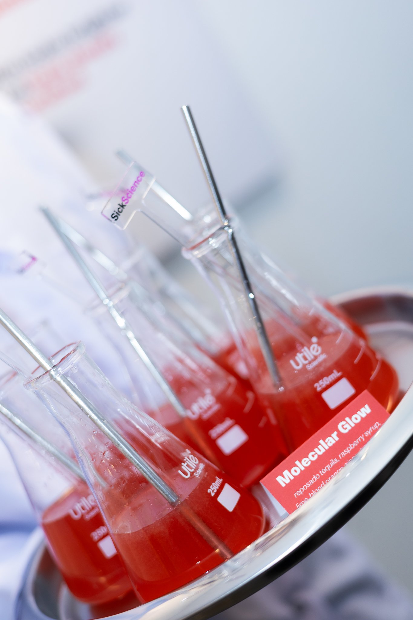 Several glass bottles filled with red liquid, labeled 'Molecular Glow', with metal mixing sticks inside, placed on a silver tray.