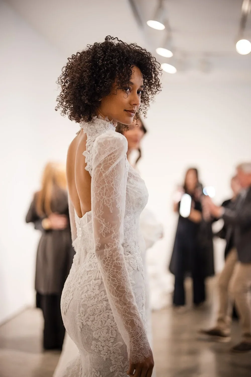 A woman in a white lace wedding dress with long sleeves, standing indoors, looking over her shoulder with a slight smile, with blurred people in the background.