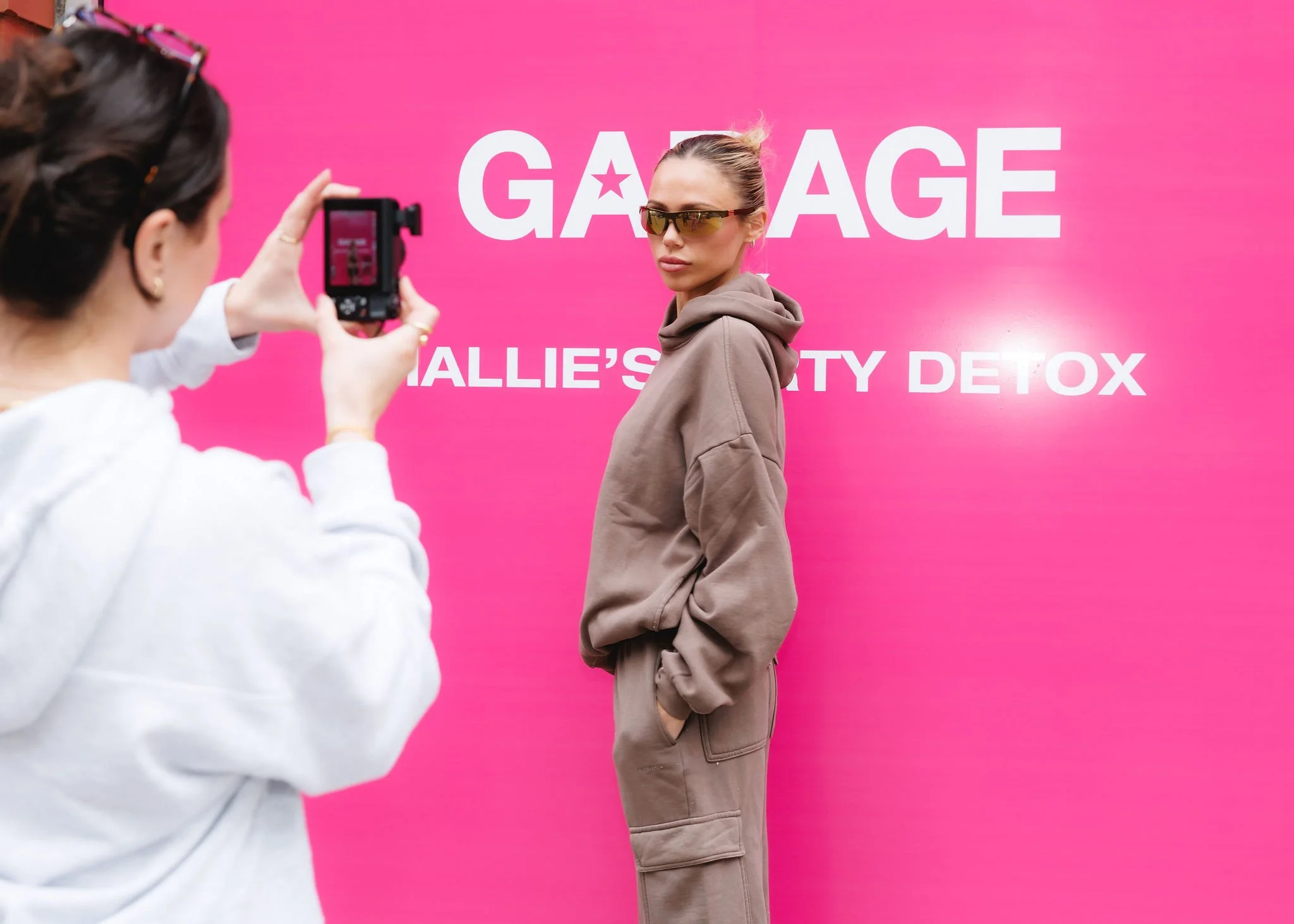 Woman in beige hoodie and cargo pants posing for photo at pink backdrop that says 'Garage Hallie's Beauty Detox' with another woman taking picture.