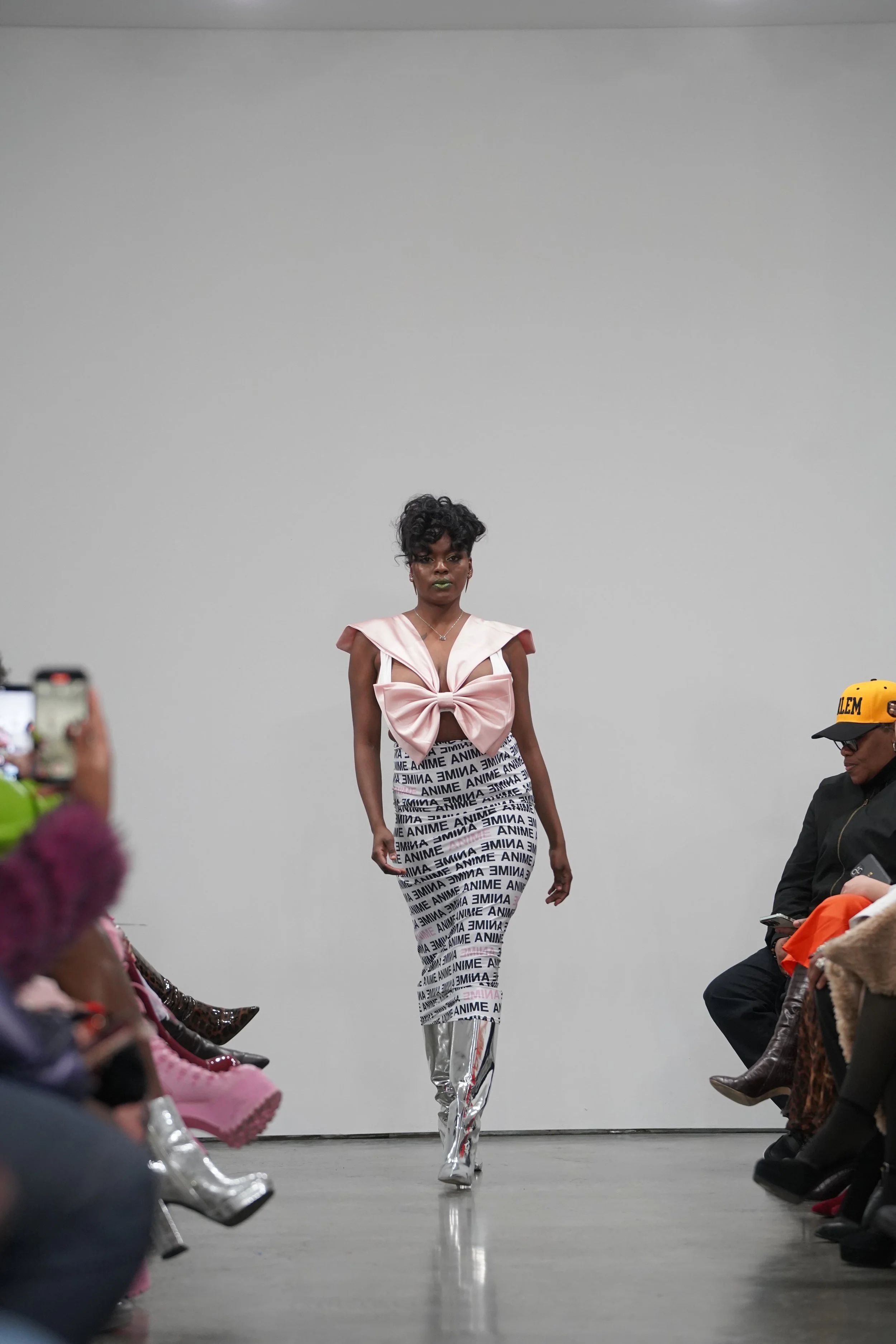 A model walking down the runway during a fashion show, wearing a pink top with a large bow and high-waisted pants with the word 'ANIME' printed repeatedly.