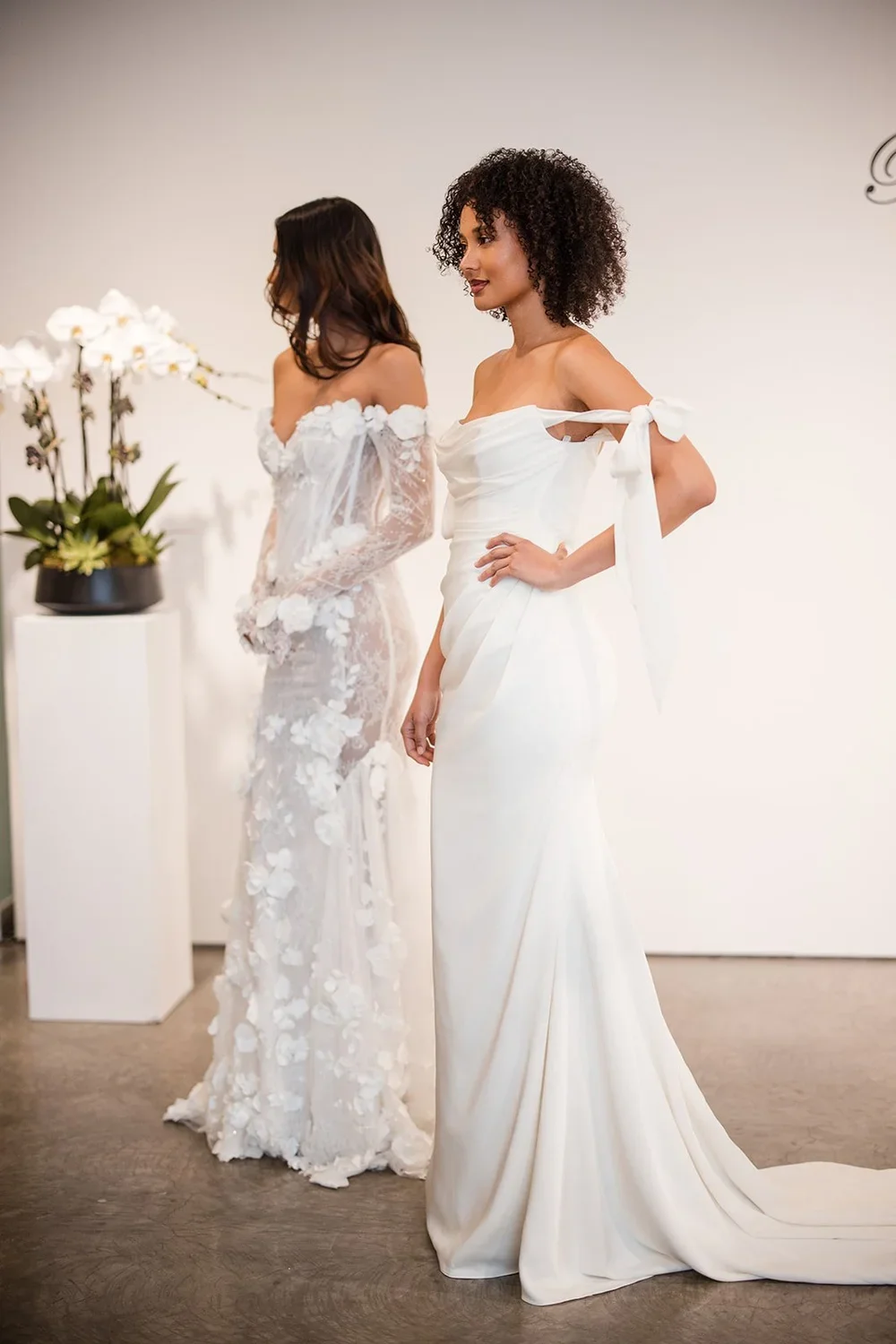 Two women in white wedding dresses standing indoors with a minimalist background, a white wall, and a potted orchid plant to the side.