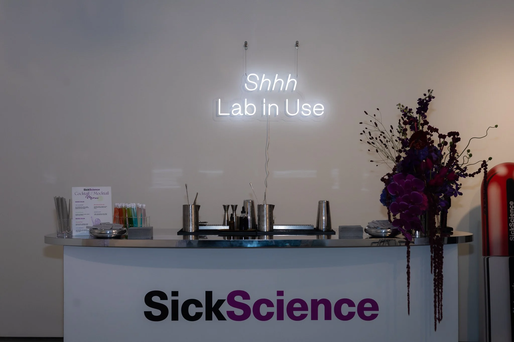 A white bar with the logo 'Sick Science' partly visible at the bottom, a neon sign on the wall above saying 'Shhh Lab in Use', and a floral arrangement on the right side of the bar. On the left side of the bar, there is a cocktail/mocktail menu, colo