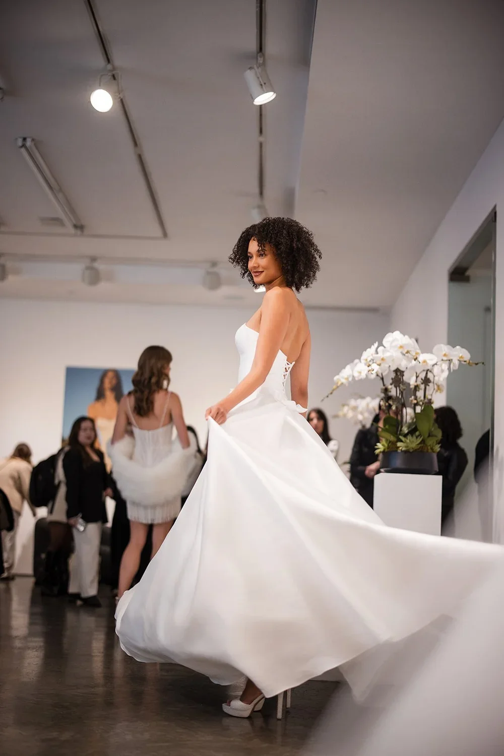 A woman modeling a white wedding dress at a bridal boutique, with a group of women and staff in the background and a vase of white orchids on the side.