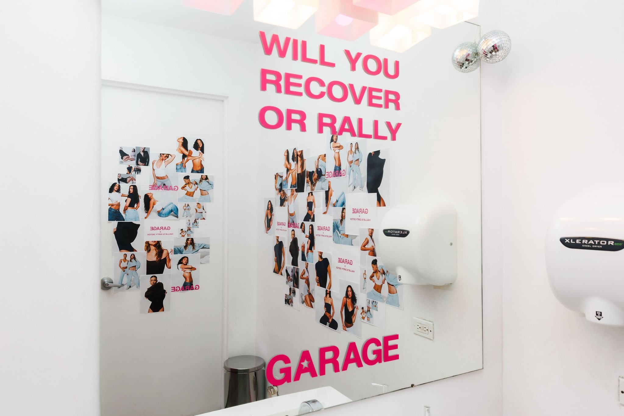 A mirror in a garage with a collage of photos of women in various outfits. Above, pink 3D letters ask, 'WILL YOU RECOVER OR RALLY' and at the bottom, 'GARAGE' with a star in the letter 'A'.