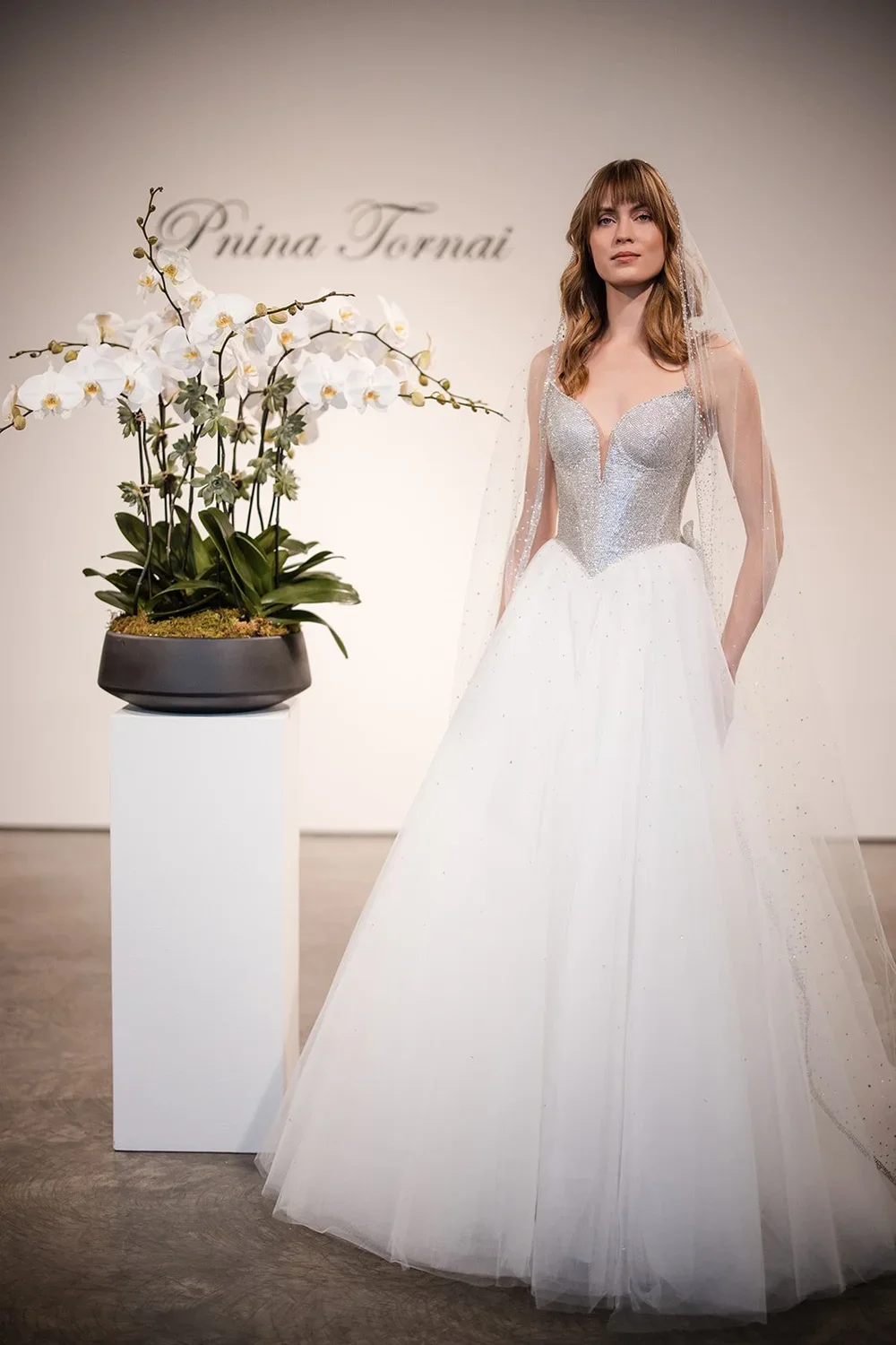 A woman in a white wedding dress with a silver bodice and long veil, standing next to a large potted white orchid plant, in an indoor setting with a plain backdrop that has the text 'Ruina Tormai'.
