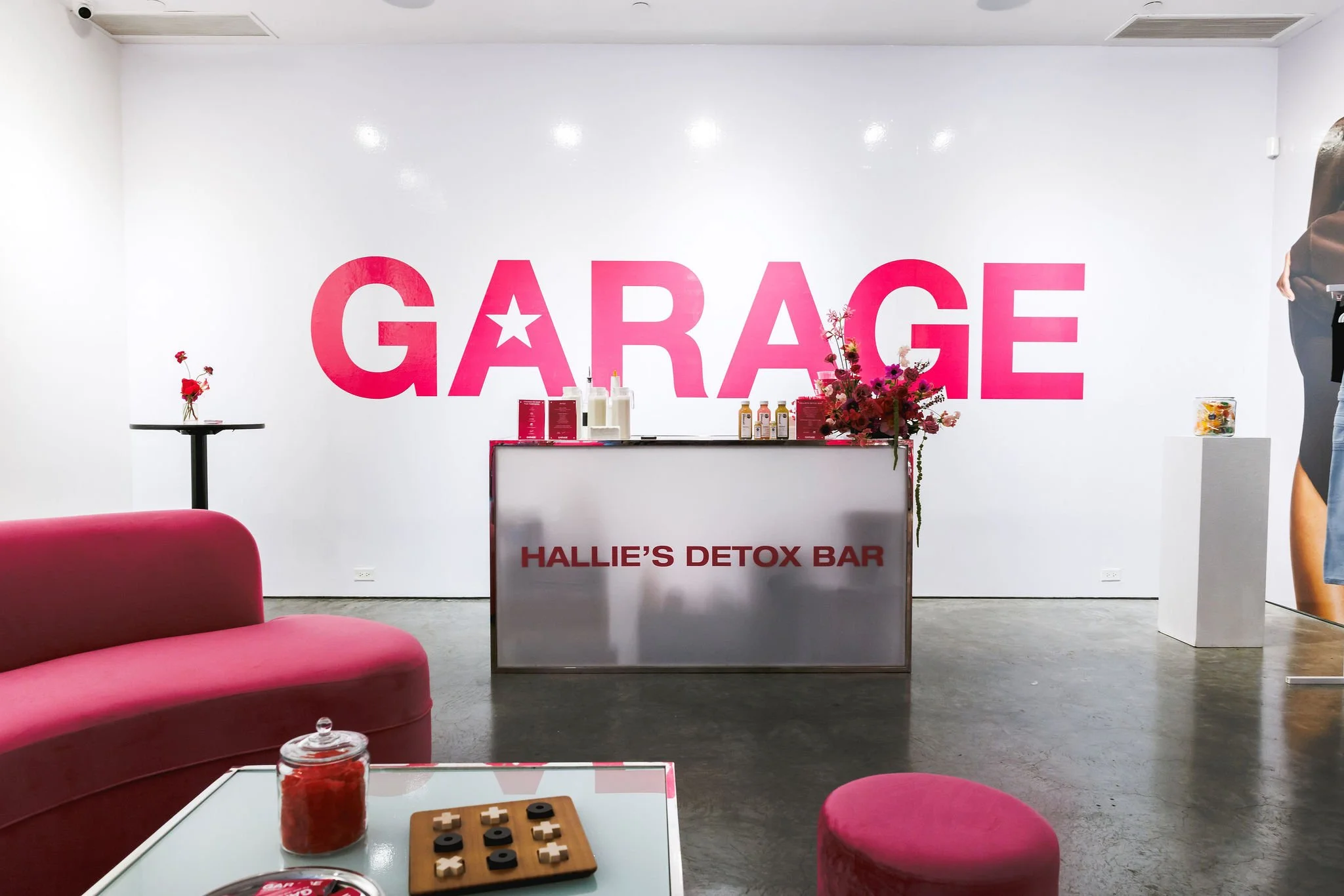 Interior of a detox bar with a white wall displaying large pink letters spelling 'GARAGE' and a star in the 'A'. Below, a reception desk with the words 'HALLIE'S DETOX BAR' in red. There is a pink sofa and a glass coffee table with a board game, a container of red candies, and snacks. To the left, a small black table with a red flower. To the right, a stand with flowers and a person partially visible.