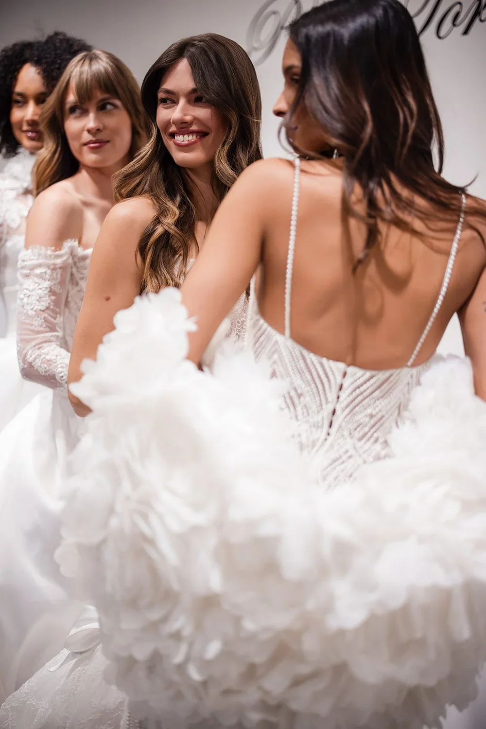 Four women in white wedding dresses standing in line, smiling and looking at each other, at a wedding event.