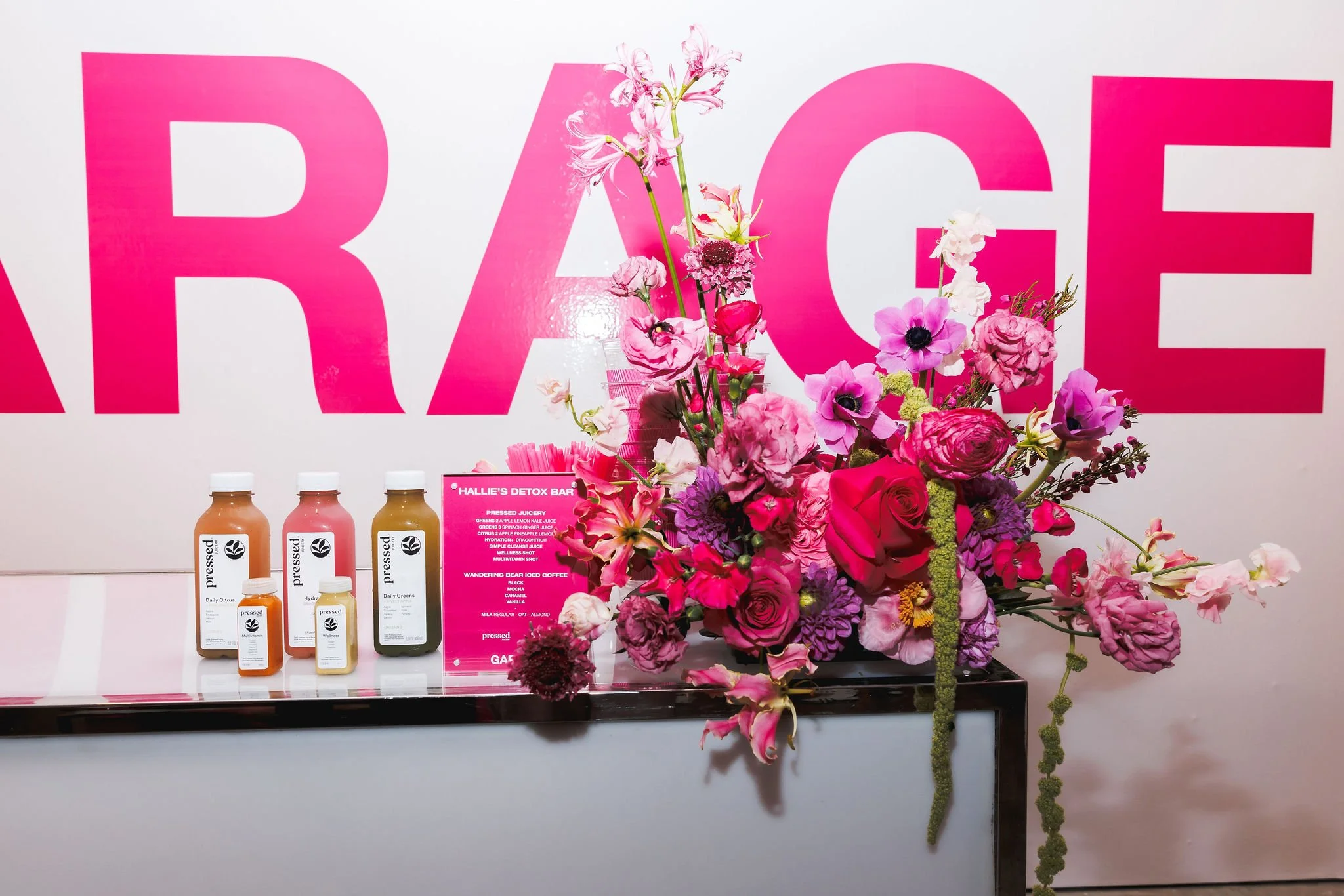 A display of bottled juices in front of large pink letters spelling 'RAGE' on a white background, with a large bouquet of pink and purple flowers, and a pink menu sign
