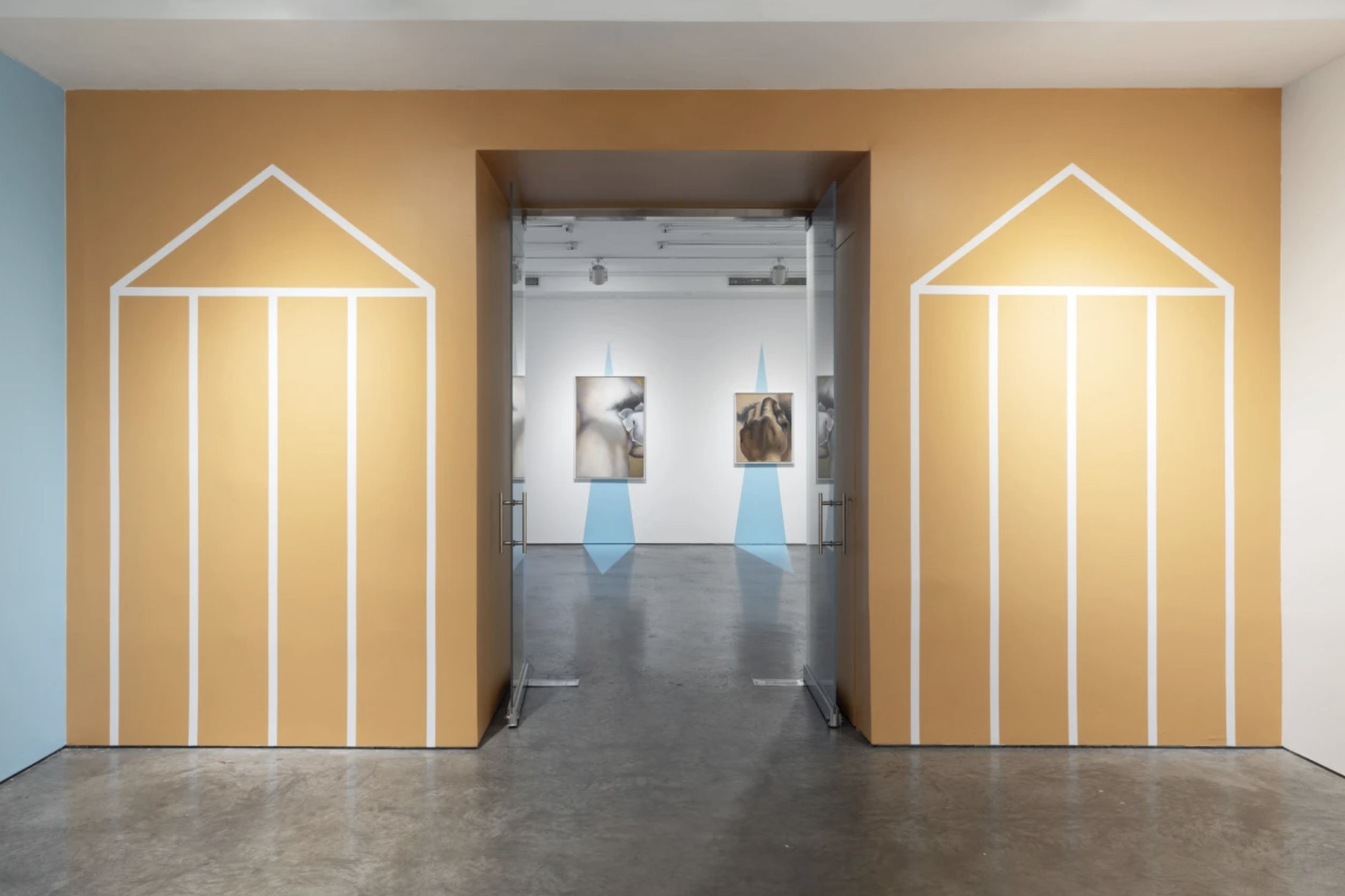 Entrance to an art gallery with orange walls decorated with white line art of a house. Inside, paintings are lit by ceiling lights and blue spotlights.