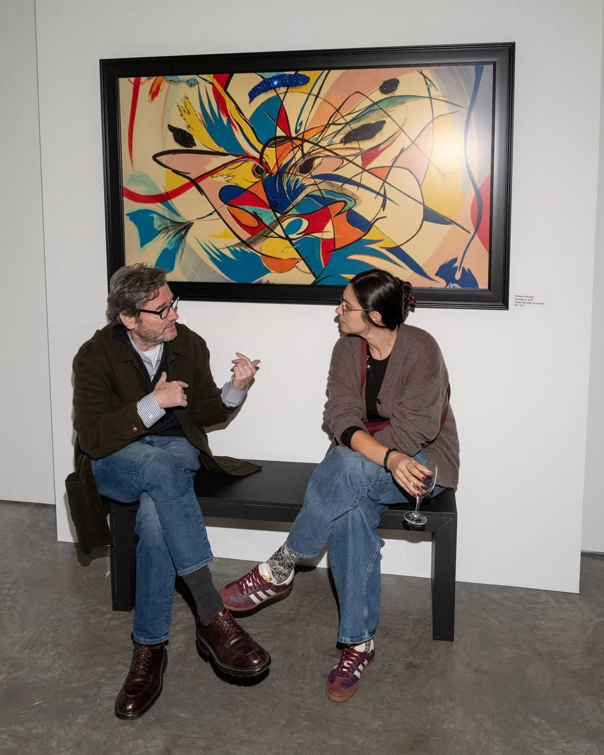 Two people sitting on a black bench, engaging in conversation at an art gallery. A large, colorful abstract painting hangs behind them on a white wall.