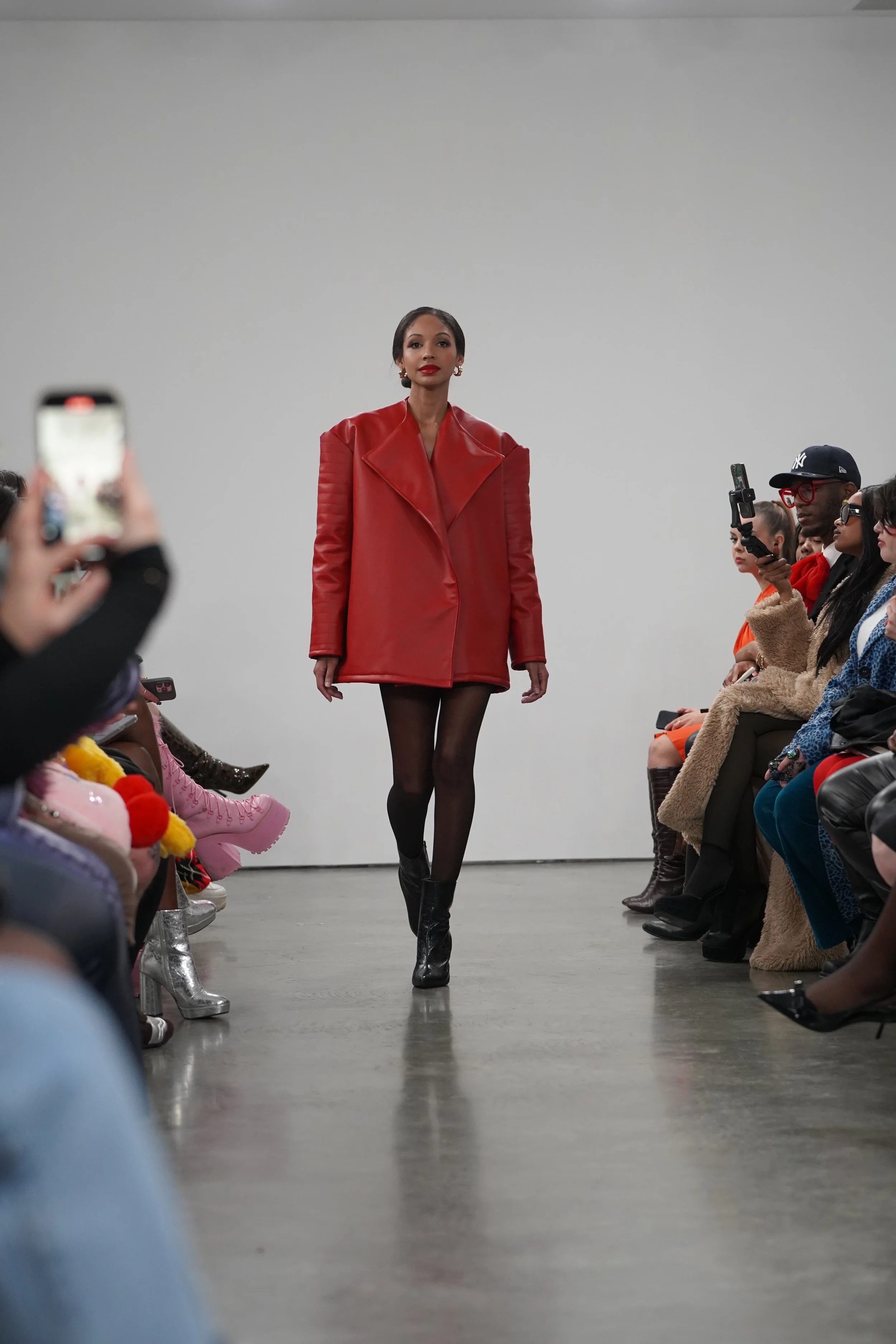 A model walking down the runway during a fashion show, wearing a red oversized coat, black tights, and black boots, with an audience seated on both sides capturing the moment.