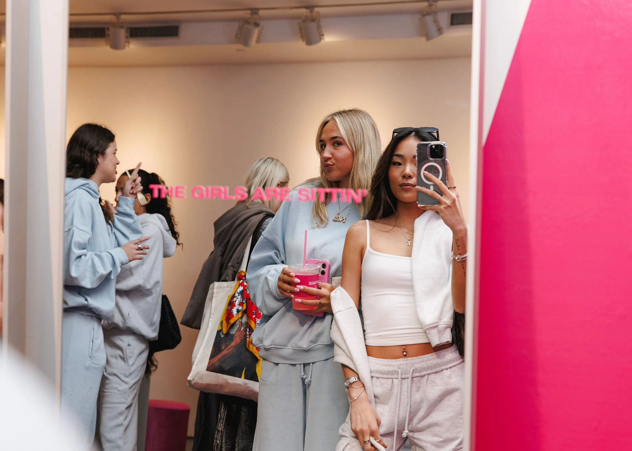 Two young women taking a selfie in front of a mirror, with others in the background. One woman wears a white crop top and beige sweatpants, holding her phone, and the other wears a grey hoodie and holds a pink drink. The mirror has pink text saying "