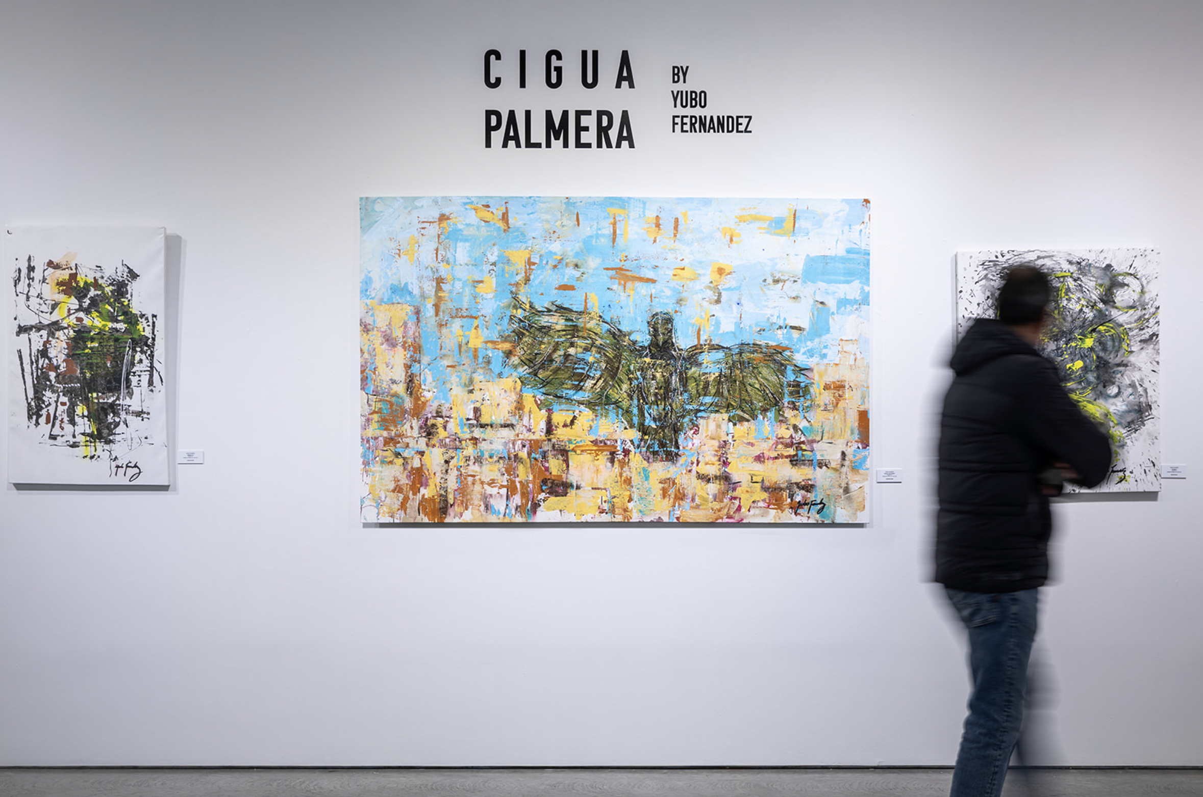 An art gallery with three abstract paintings on a white wall and a person in a black jacket looking at the artwork.