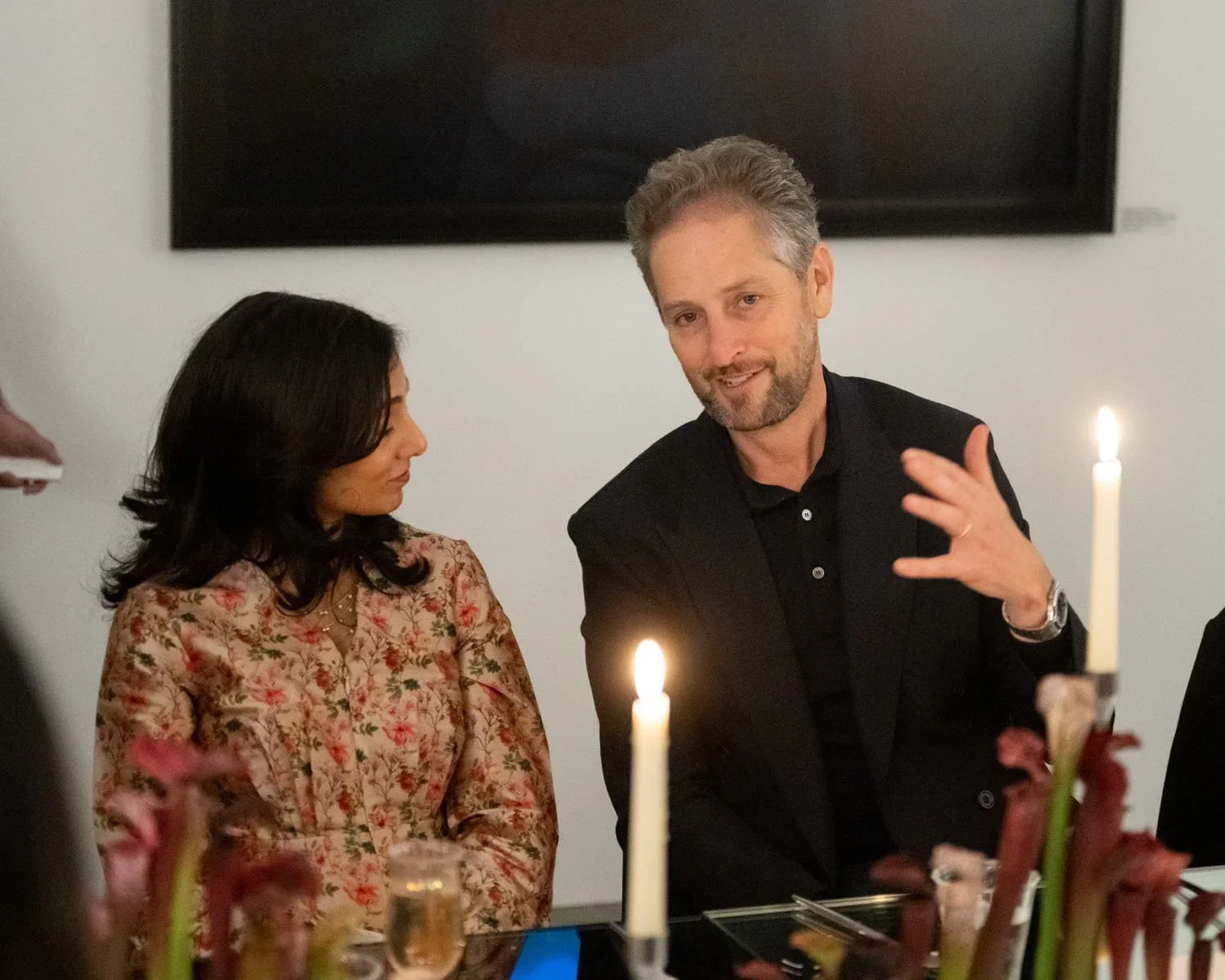 A man with gray hair and beard wearing a black suit and shirt, gesturing with his right hand while speaking. A woman with dark hair wearing a floral blouse looks at him. The setting appears to be a dinner or social gathering with candles and wine gla