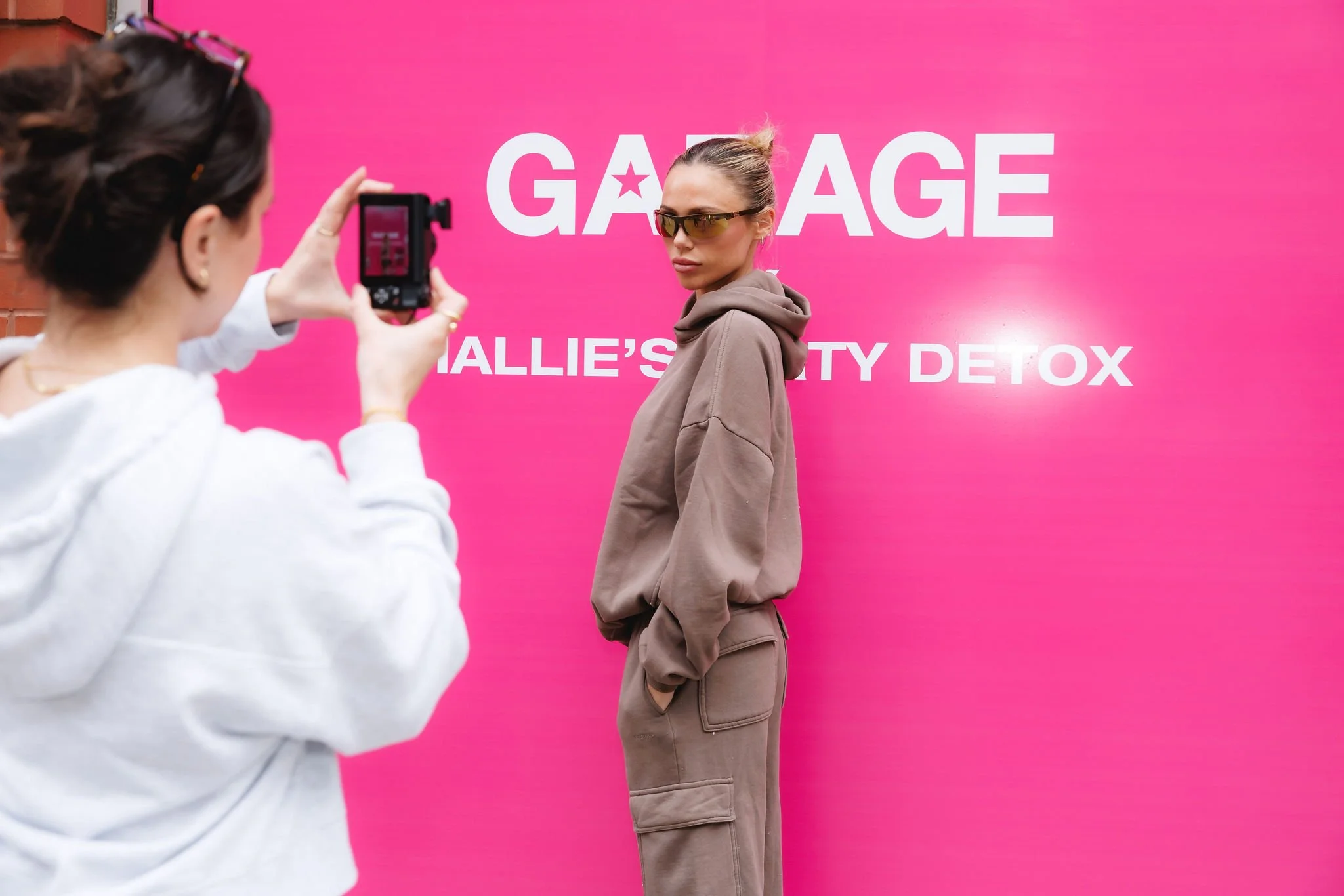 A woman in a brown hoodie and pants poses in front of a pink wall with large white text that reads 'GARAGE ALLIE'S BEAUTY DETOX'. Another person takes her photo with a camera.