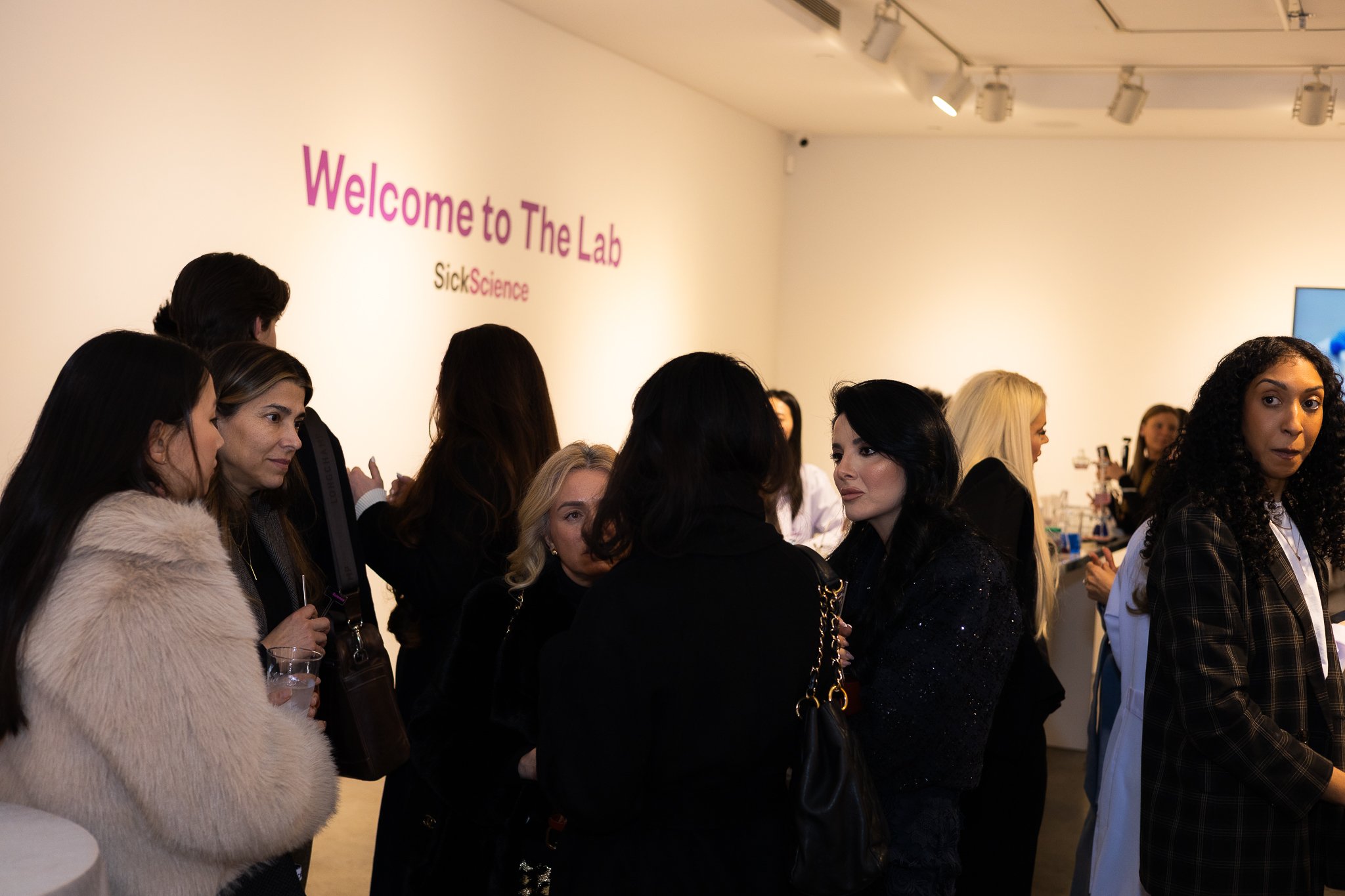 A group of women talking and socializing in a room with a sign that says "Welcome to The Lab" and "Sick Science" on the wall.