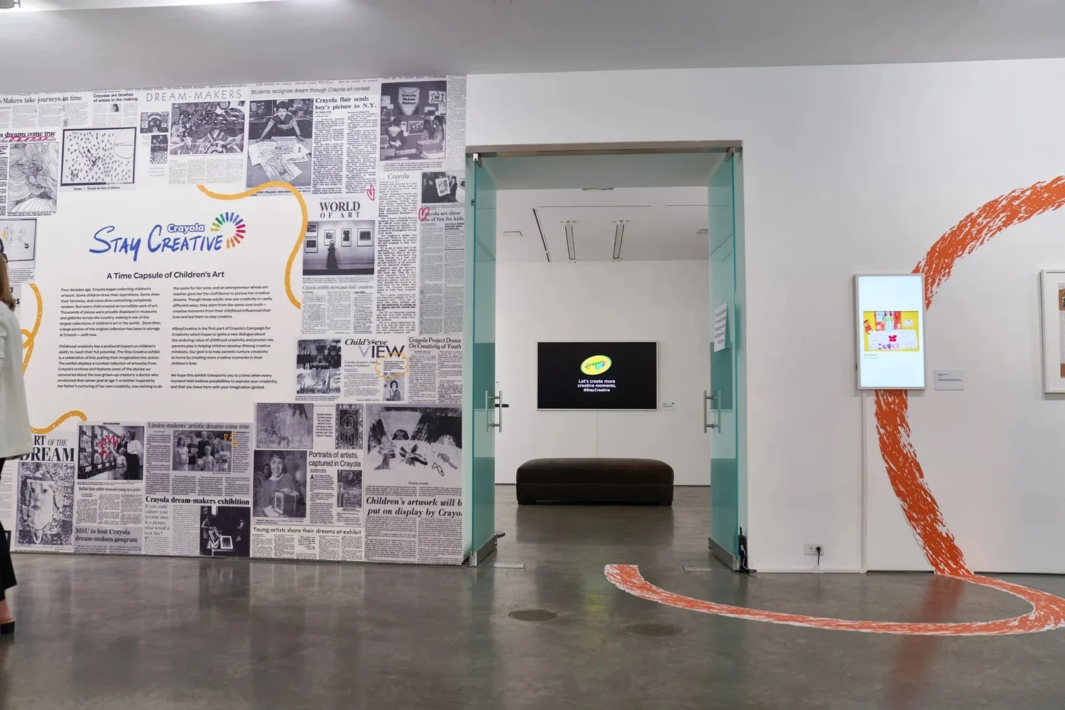 An art exhibit titled 'Stay Creative' featuring a wall decorated with newspaper clippings about children's art, a digital display with a campaign message, and a curved orange line painted on the white wall leading to a doorway. Inside the room, a bla