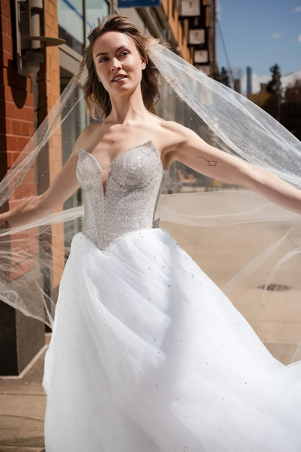 A woman in a wedding dress with a beaded bodice and a flowing white skirt, outdoors on a city street, with her arms outstretched and a veil flowing behind her.