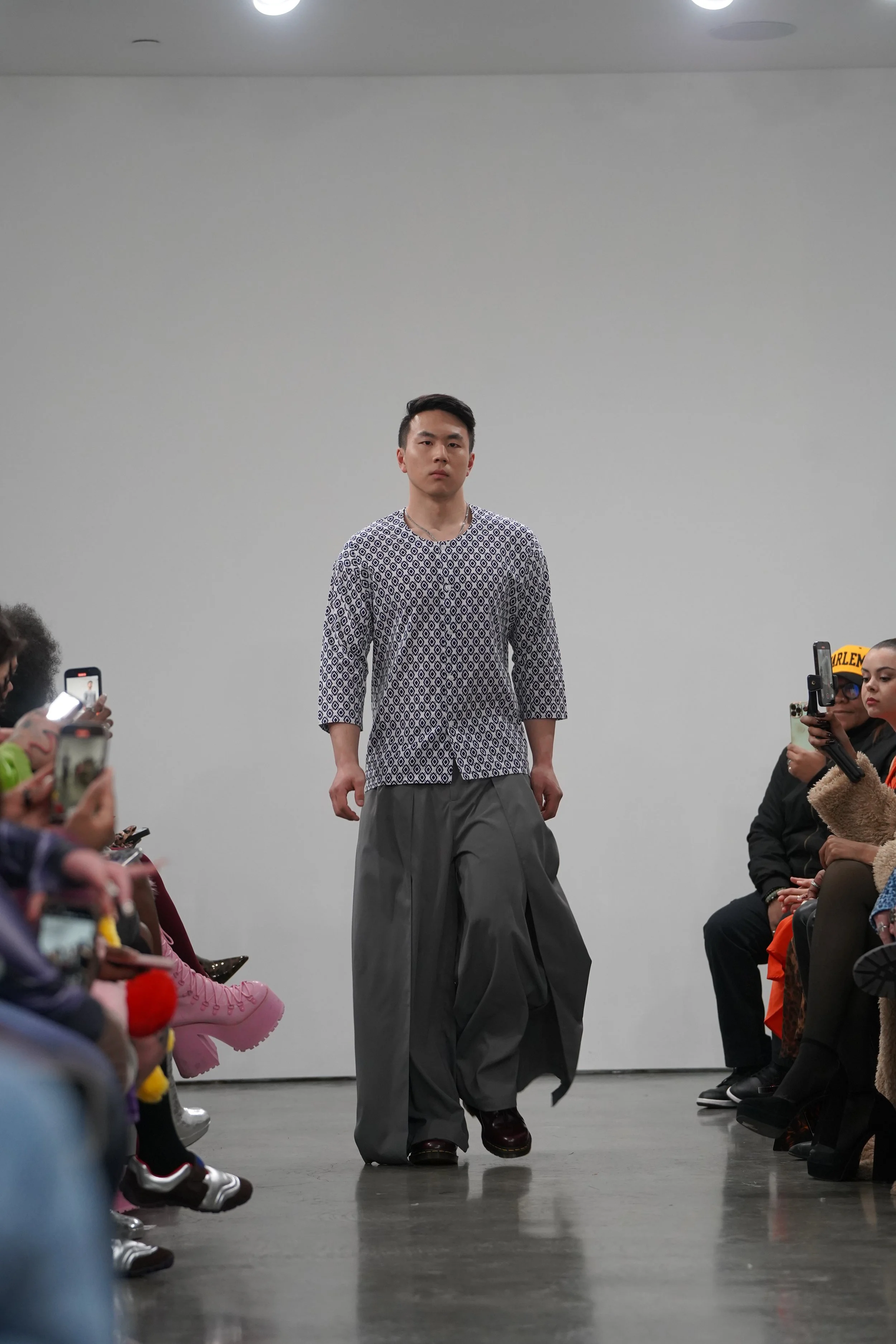 Model walking down runway at a fashion show, wearing patterned top and oversized gray pants, with audience taking photos.