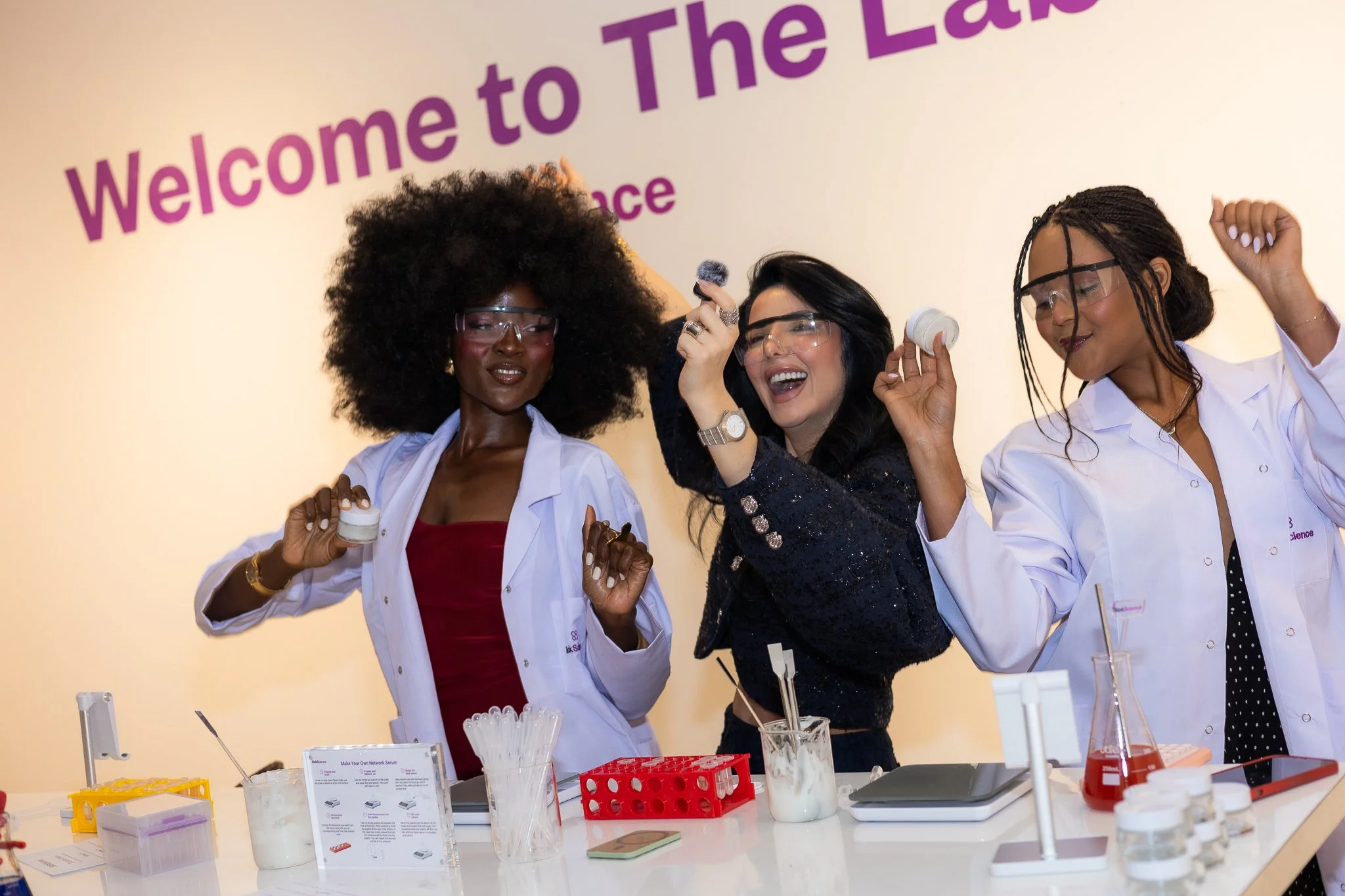 Three women, two dressed in white lab coats and one in a black dress, celebrating with laboratory equipment on a table in front of them. They are smiling and holding small containers, with a "Welcome to The Lab" sign in the background.