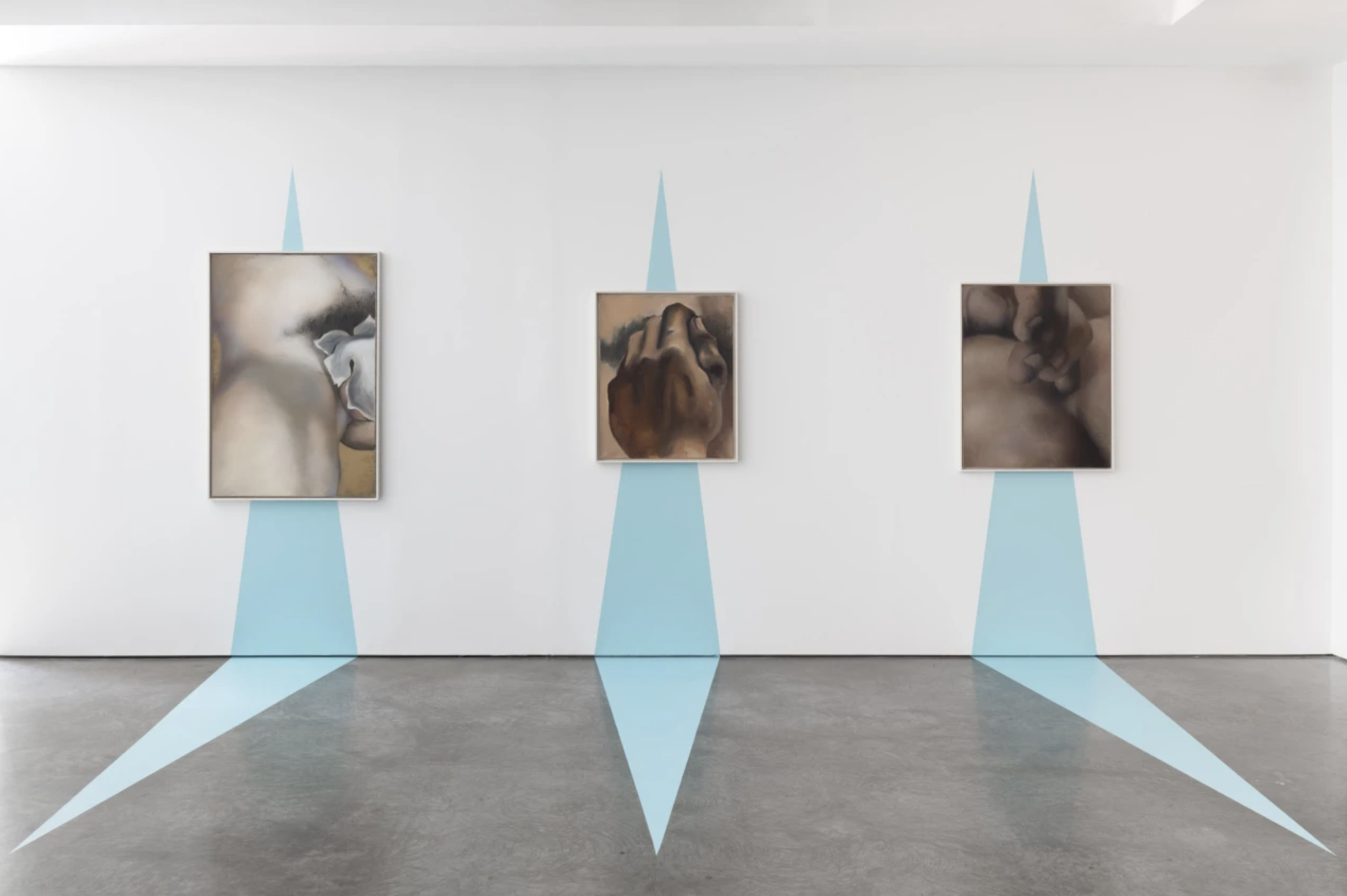 Art gallery with three framed paintings on a white wall, side-by-side. Light blue geometric shapes on the wall and floor create a symmetrical pattern.