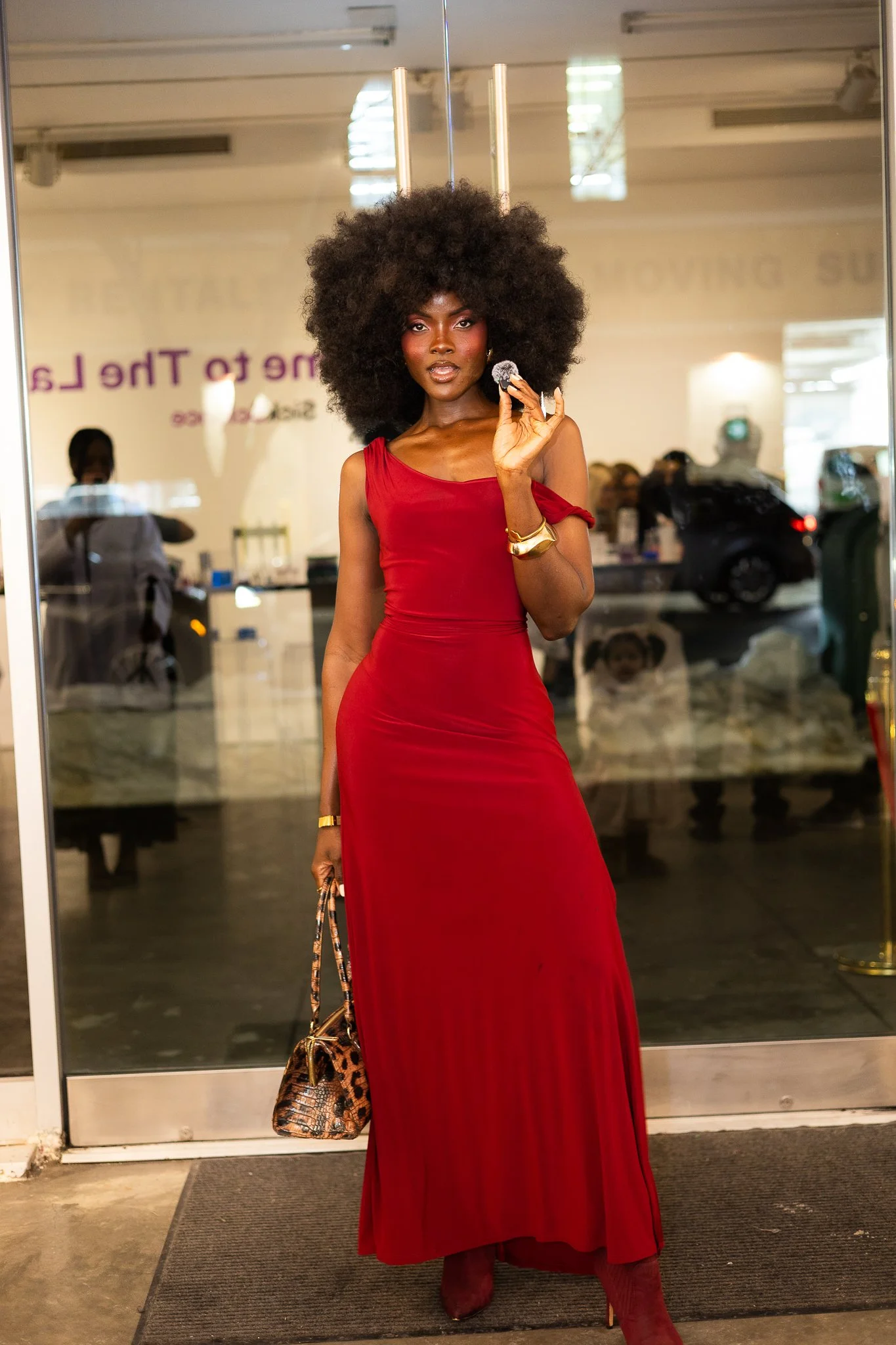A woman with dark skin and a large curly afro hairstyle is wearing a long red dress and maroon heels. She is standing in front of a glass door, holding a small object in her right hand and a leopard print handbag in her left hand, with a gold bracele