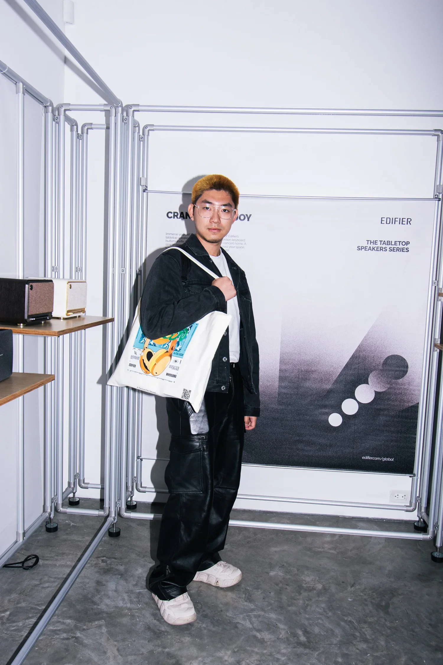 Young man with short hair and glasses standing in front of a display booth at an event, holding a white tote bag with colorful artwork, wearing a black jacket, white shirt, black leather pants, and white sneakers.