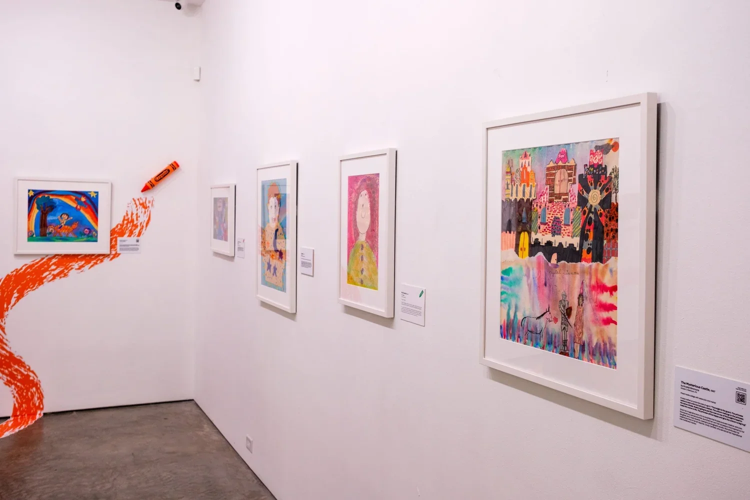 Gallery wall displaying framed colorful artwork with a focus on a vibrant castle scene and characters, textured and abstract styles, with a painted orange swirl on a white wall.